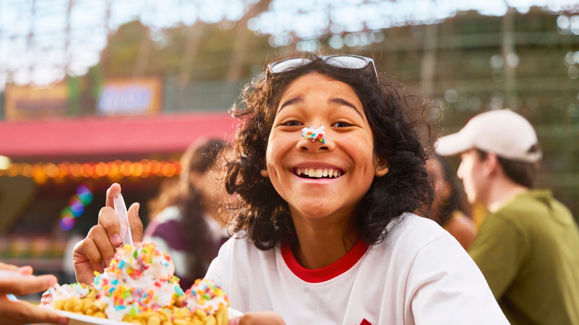 Smiling person with curly hair and sunglasses on head has colorful sprinkles on their nose, holding a plate with a large dessert topped with whipped cream and rainbow sprinkles outdoors.