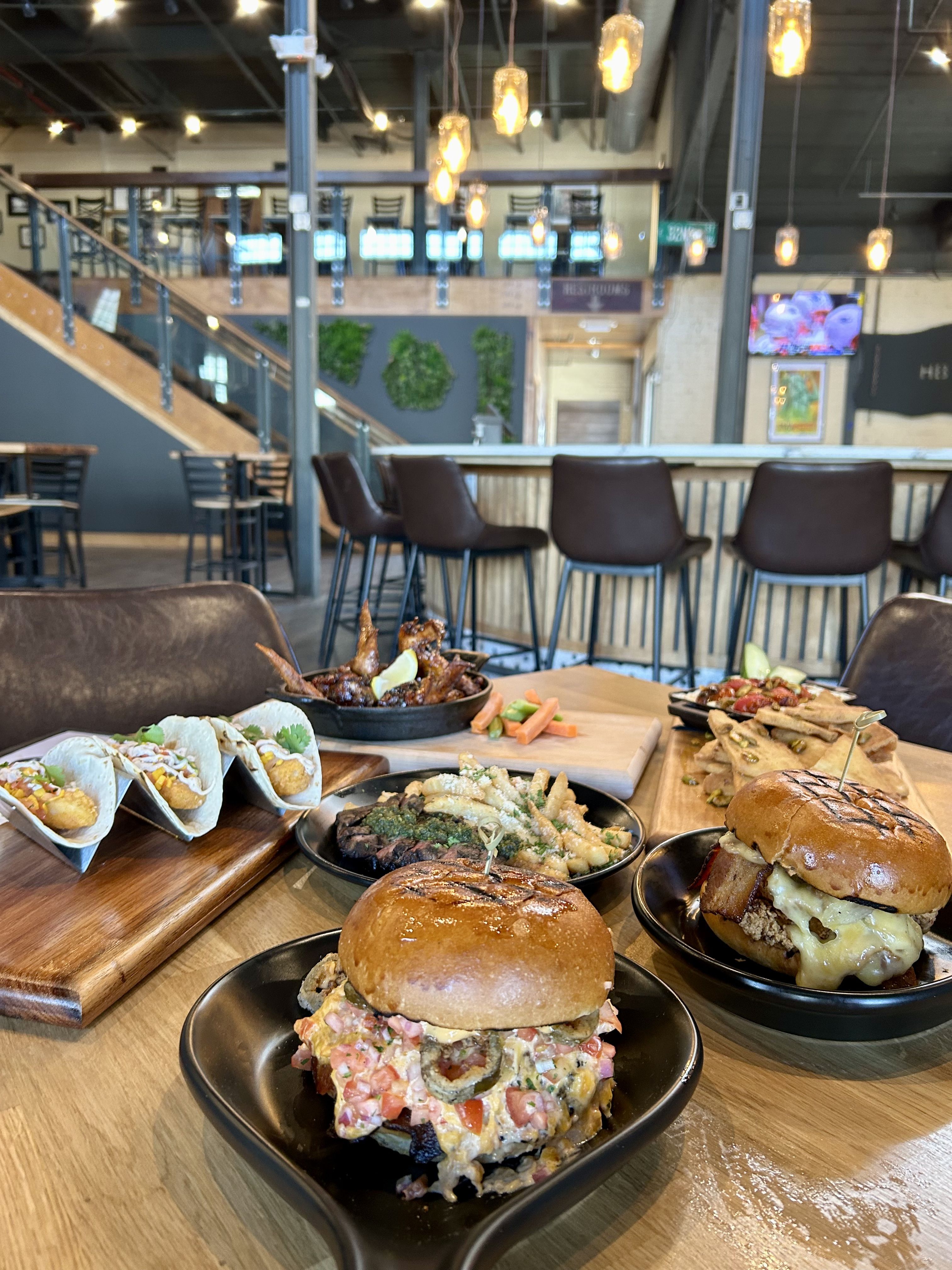 Industrial-style restaurant interior with warm hanging bulbs, metal railings and a staircase in back. Foreground shows burgers, tacos and assorted appetizers on wooden boards.