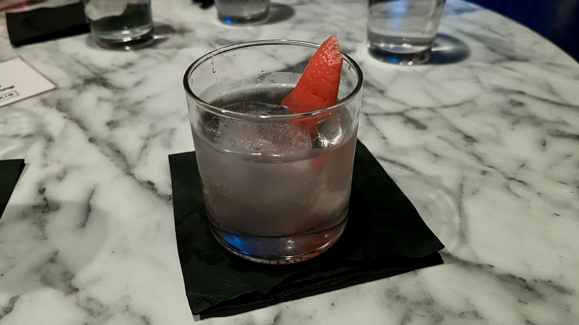 A clear cocktail with an orange twist sitting on a black and white marble table.