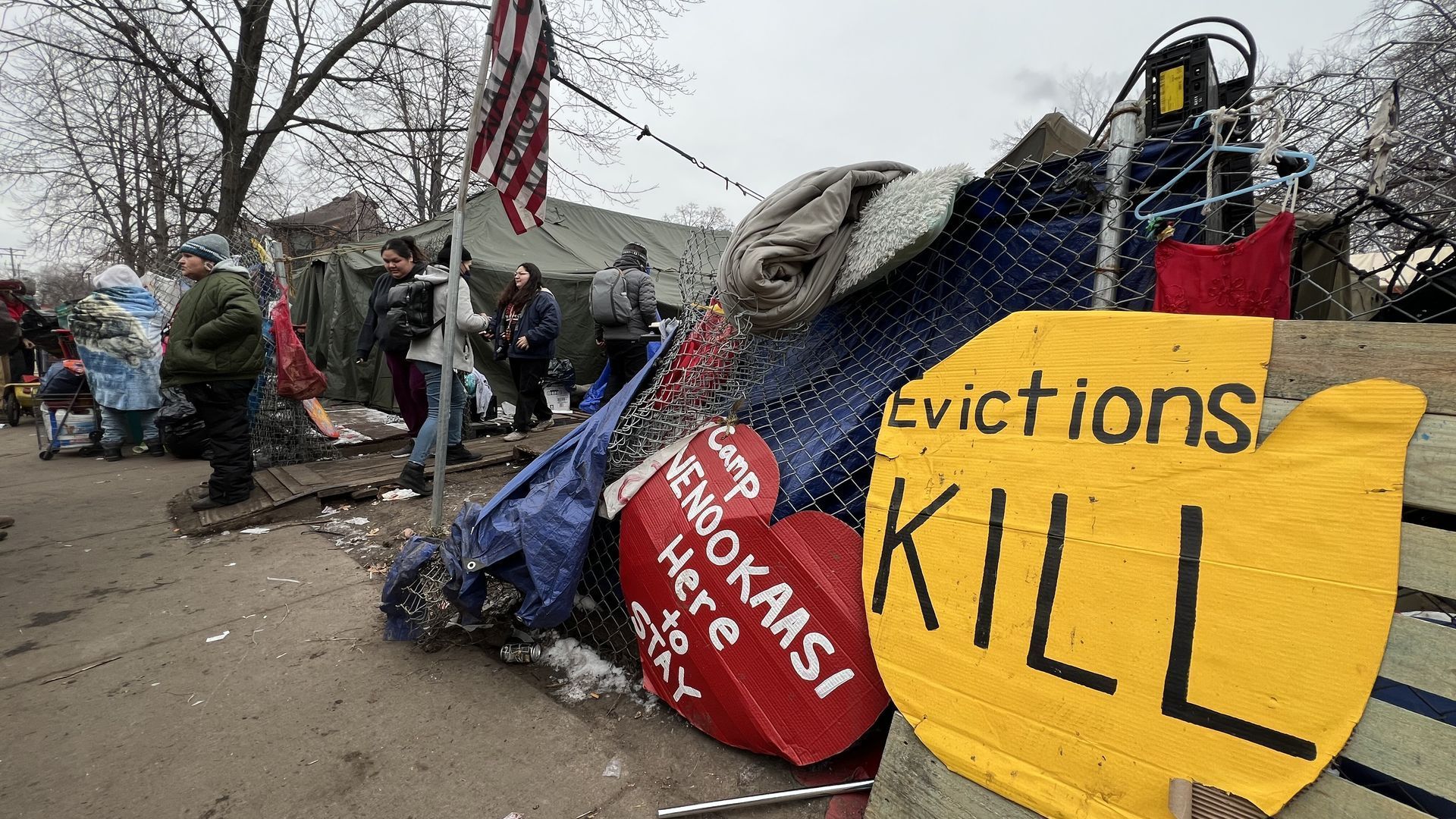 A bright yellow sign reading 'Evictions KILL' hangs on a fence outside a homeless encampment with an American flag waving over the entrance and tents visible in the background.