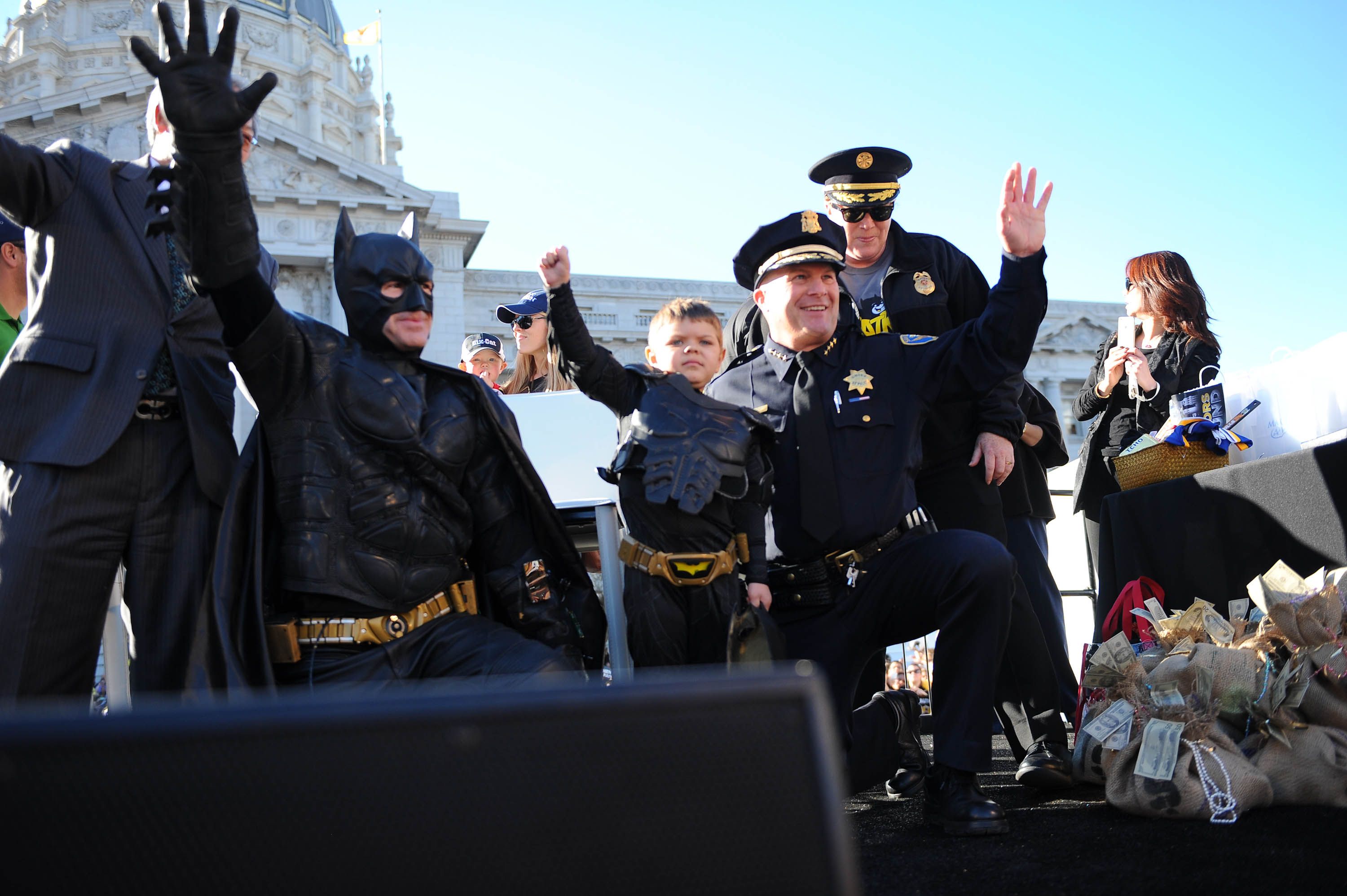 Photo of a small boy in a Batkid costume raising his arm as he poses with SFPD officers 