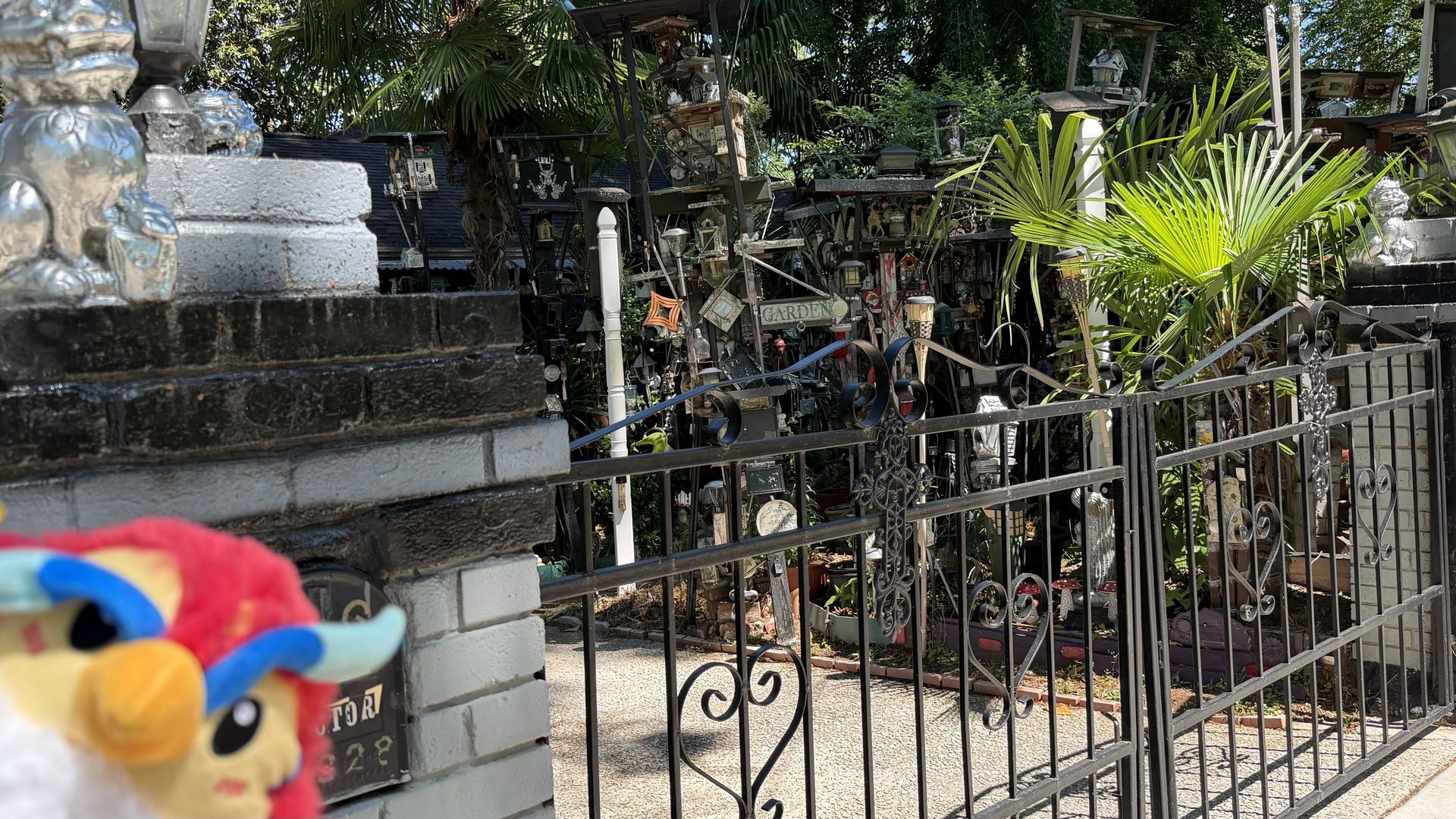 Cluttered outdoor garden yard with shelves of ornaments, statues, lanterns, and signs. A black metal fence spans the foreground; bright green palm fronds fill the right.