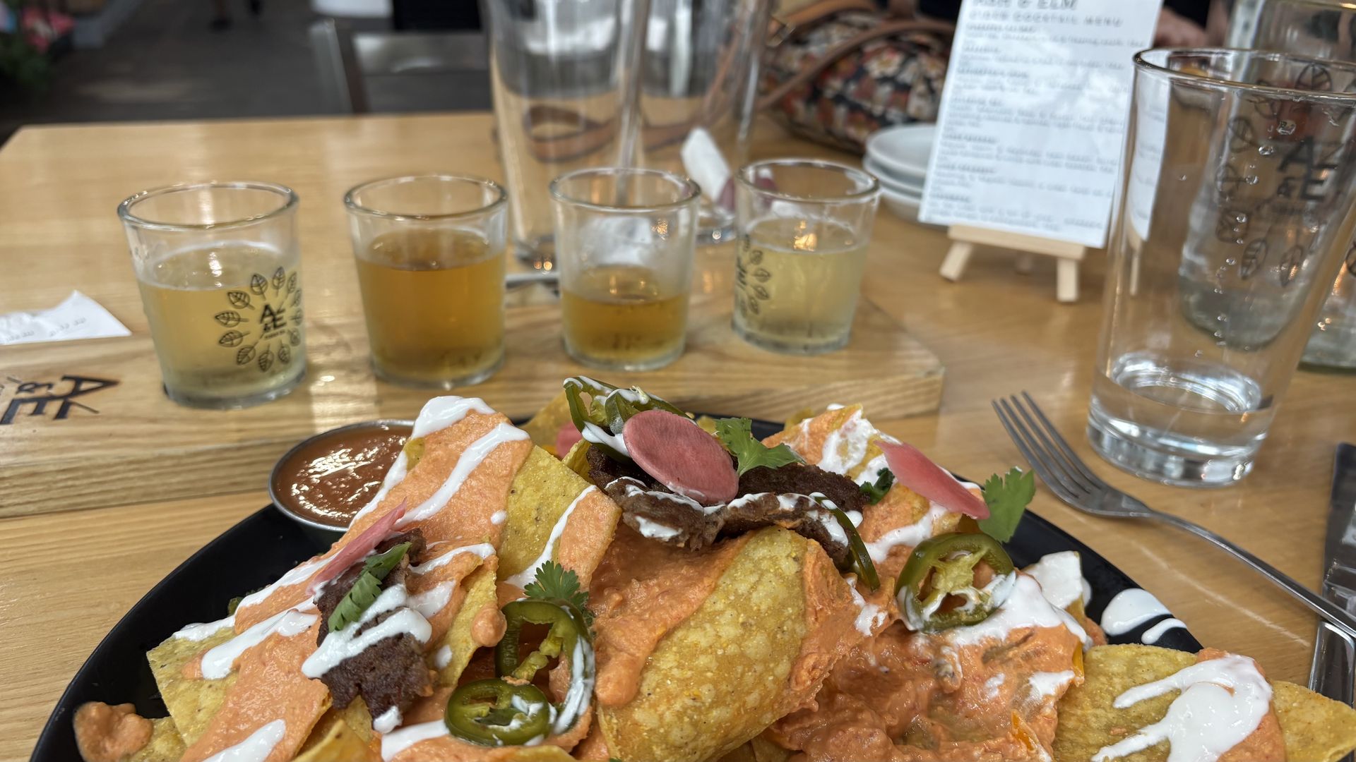 Plate of loaded nachos with cheese, jalapeños, cilantro, and pickled vegetables topped with white sauce on a wooden table. Behind, four small glasses with light drinks on a wooden flight board.