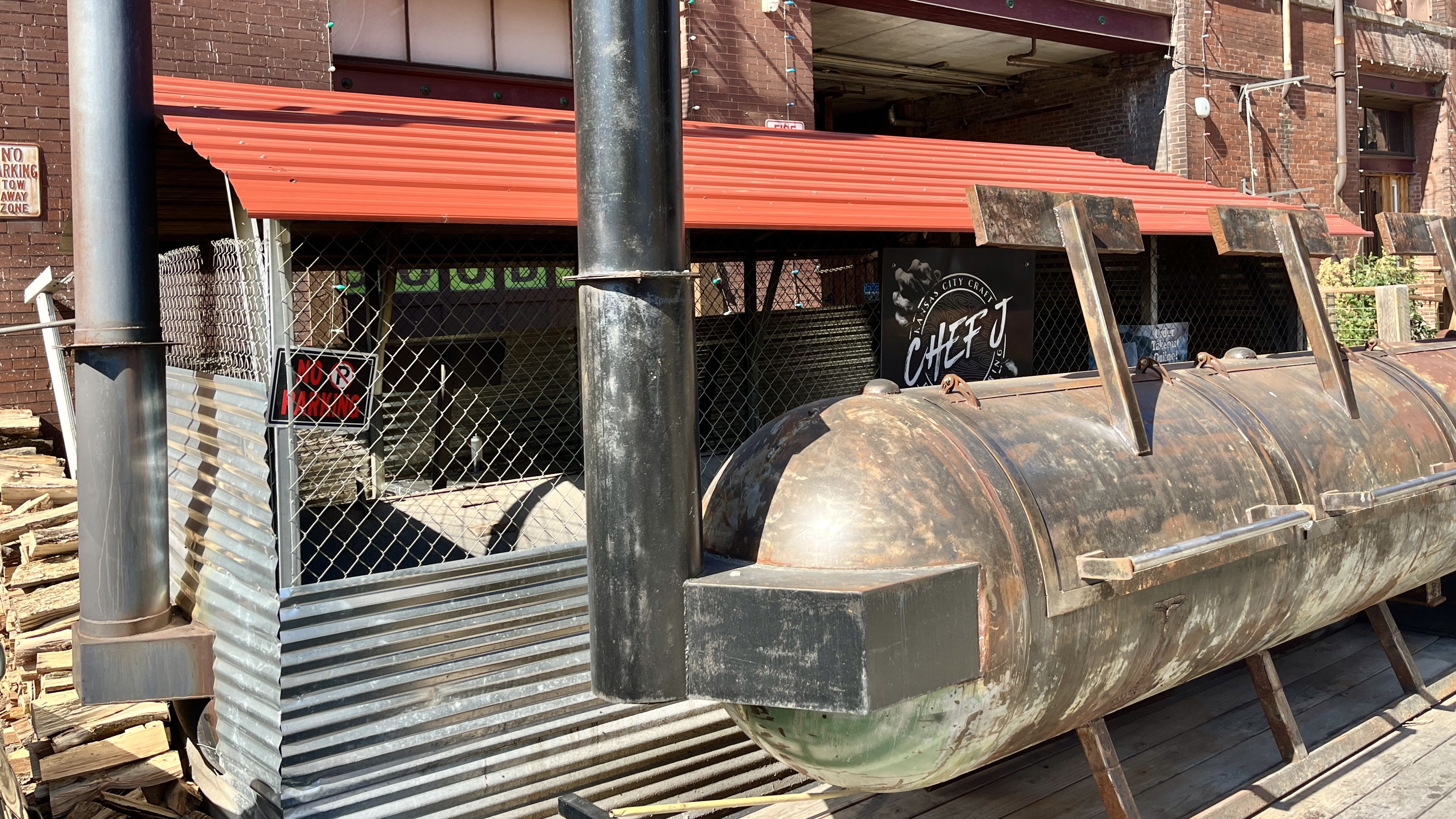 Rusty large metal smoker grill with two vertical pipes in front of a chain-link fenced area with a red corrugated roof, a pile of wood, and a sign reading "CHEF J"