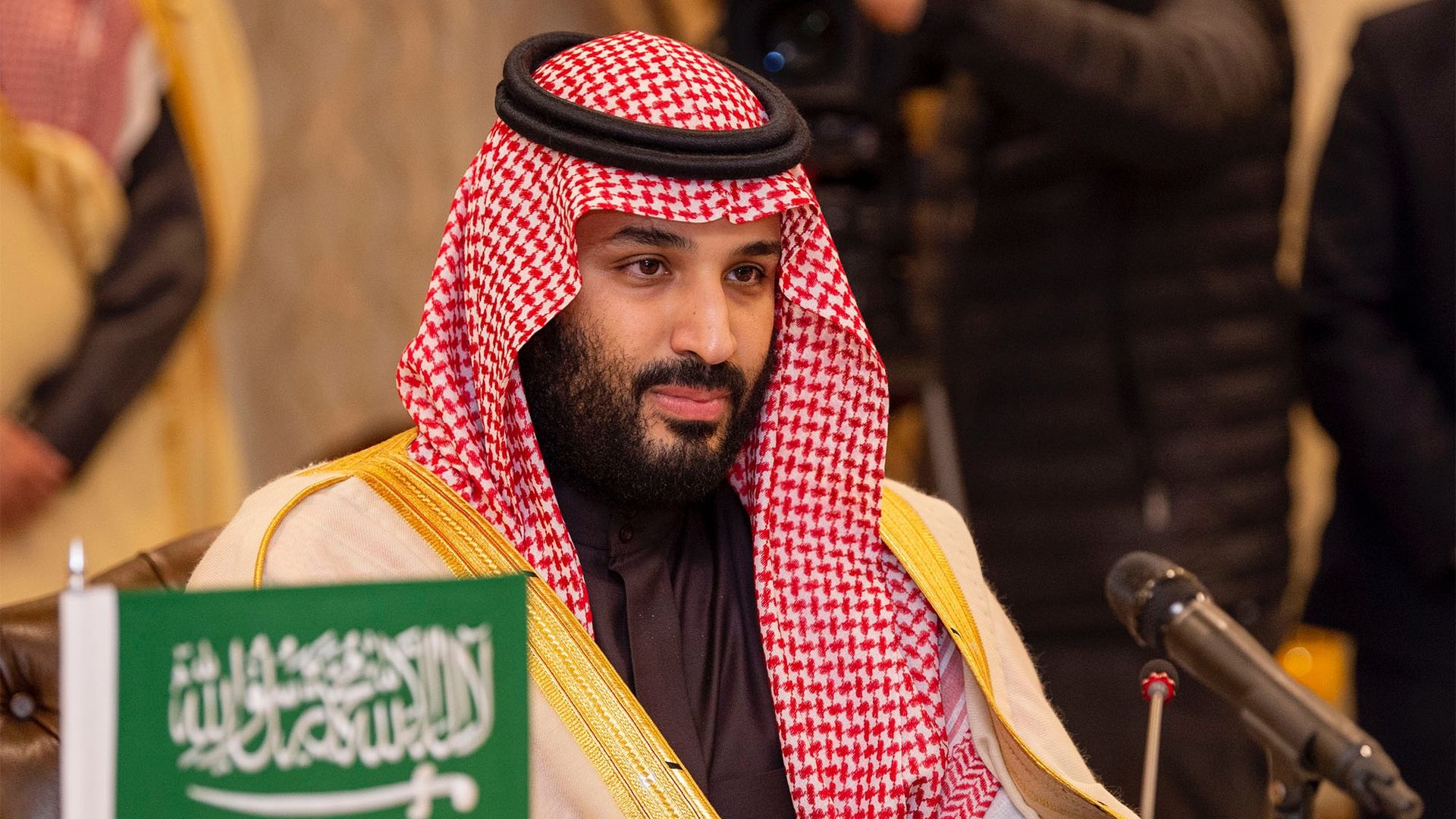 In this image, Crown Prince of Saudi Arabia Mohammad bin Salman sits behind a small green and white Saudi Arabian flag.