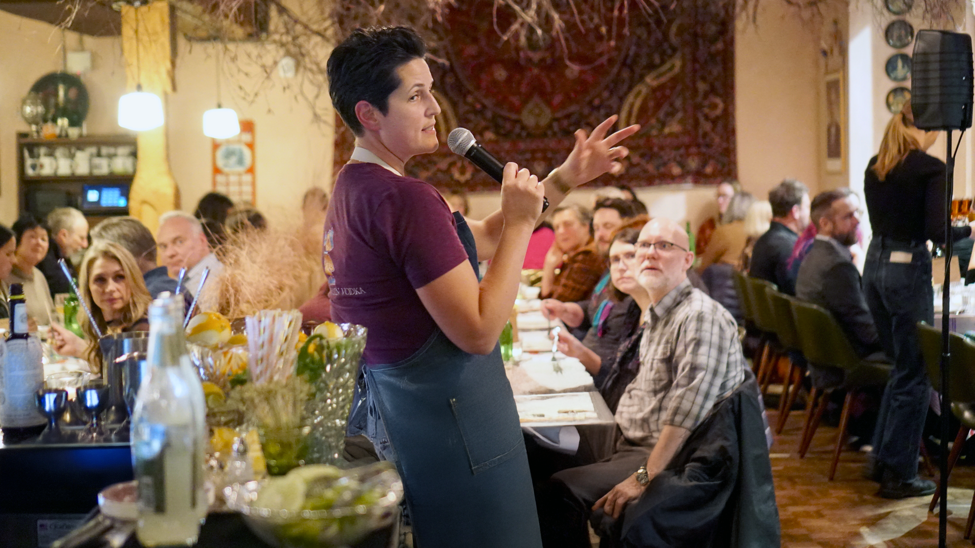 A chef speaks to diners who are seated at long tables.