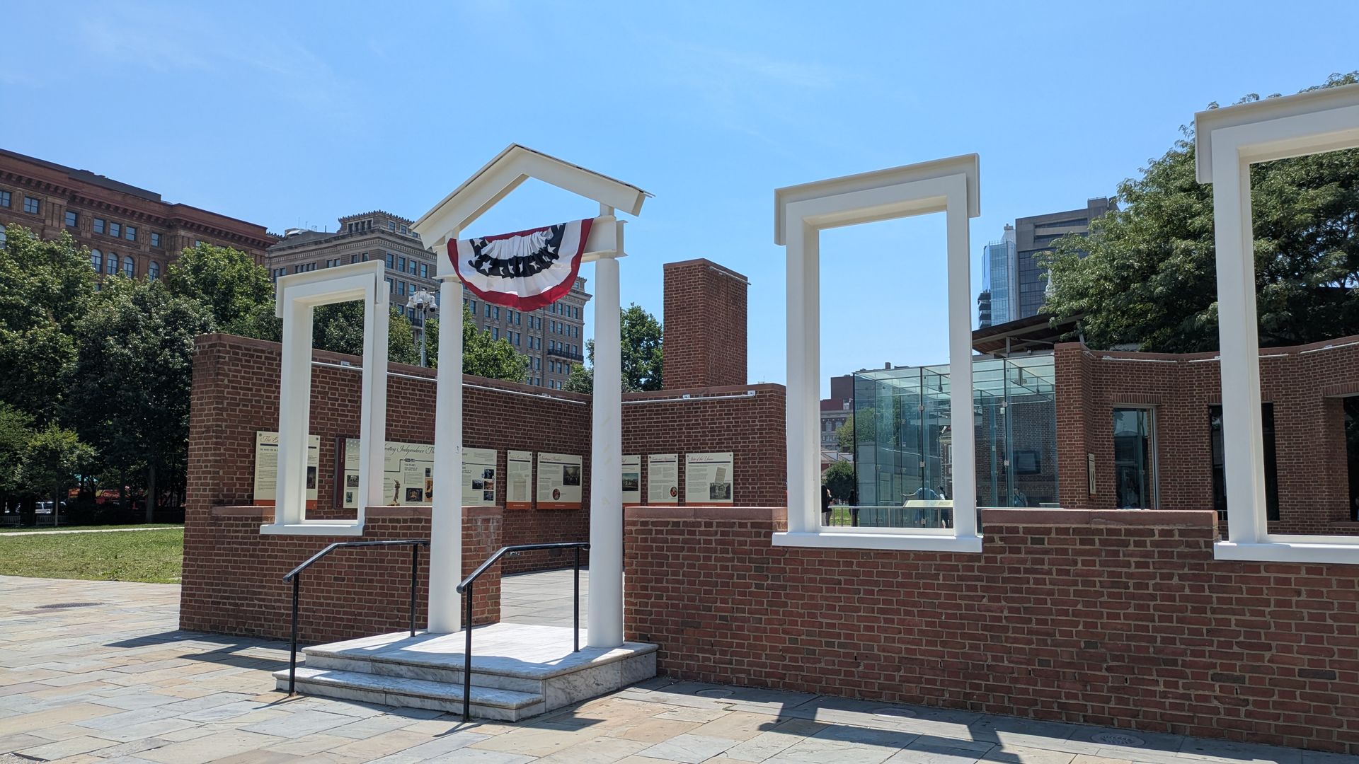 Open plaza with white rectangular arches set in a red brick wall, a red, white, and blue bunting draped on one arch, brick walls and display boards, trees and city buildings under a clear blue sky.