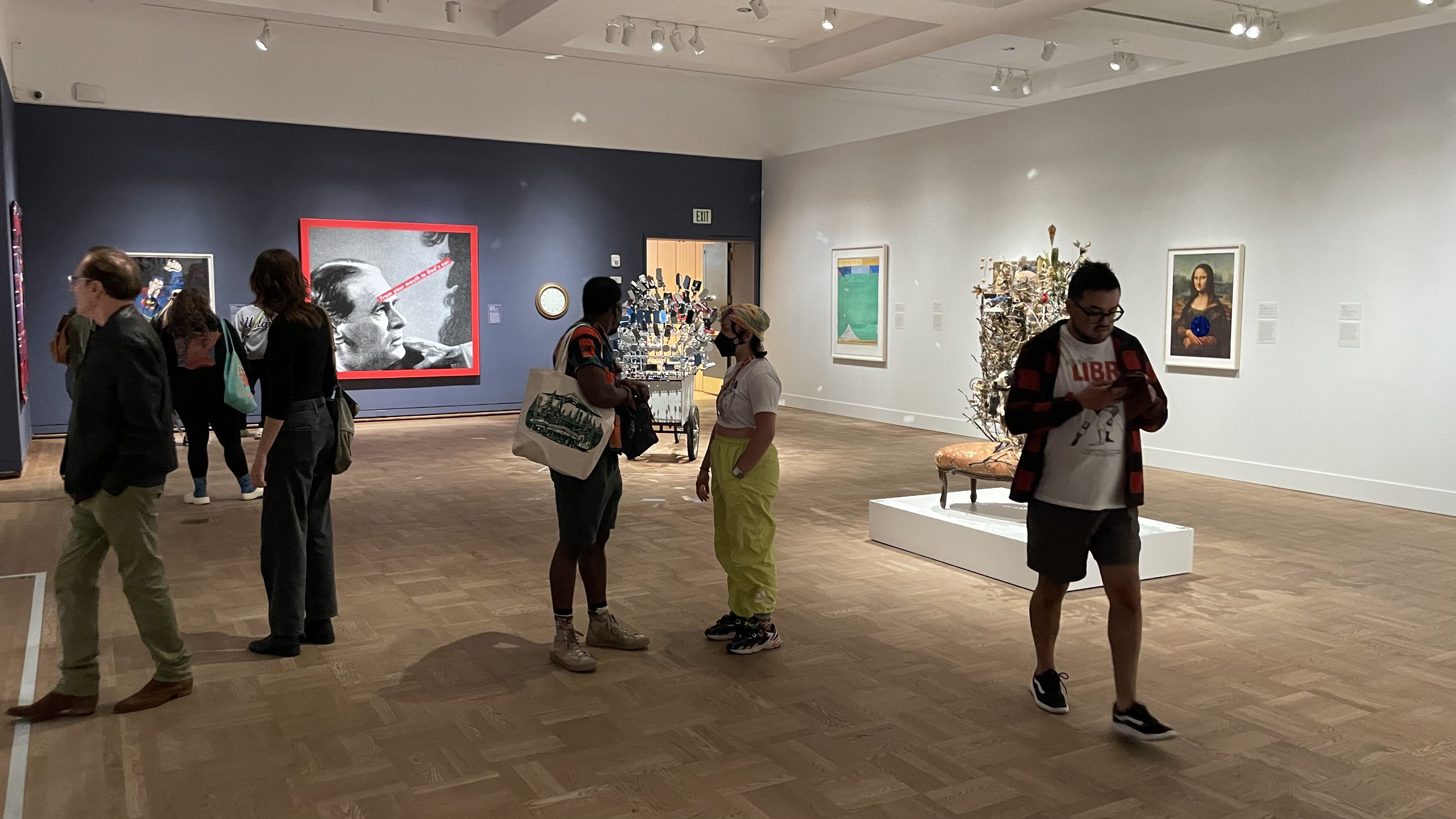 People viewing modern and classical art in a well-lit gallery with light wooden floors, including a black-and-white portrait with red border and a framed image resembling the Mona Lisa.