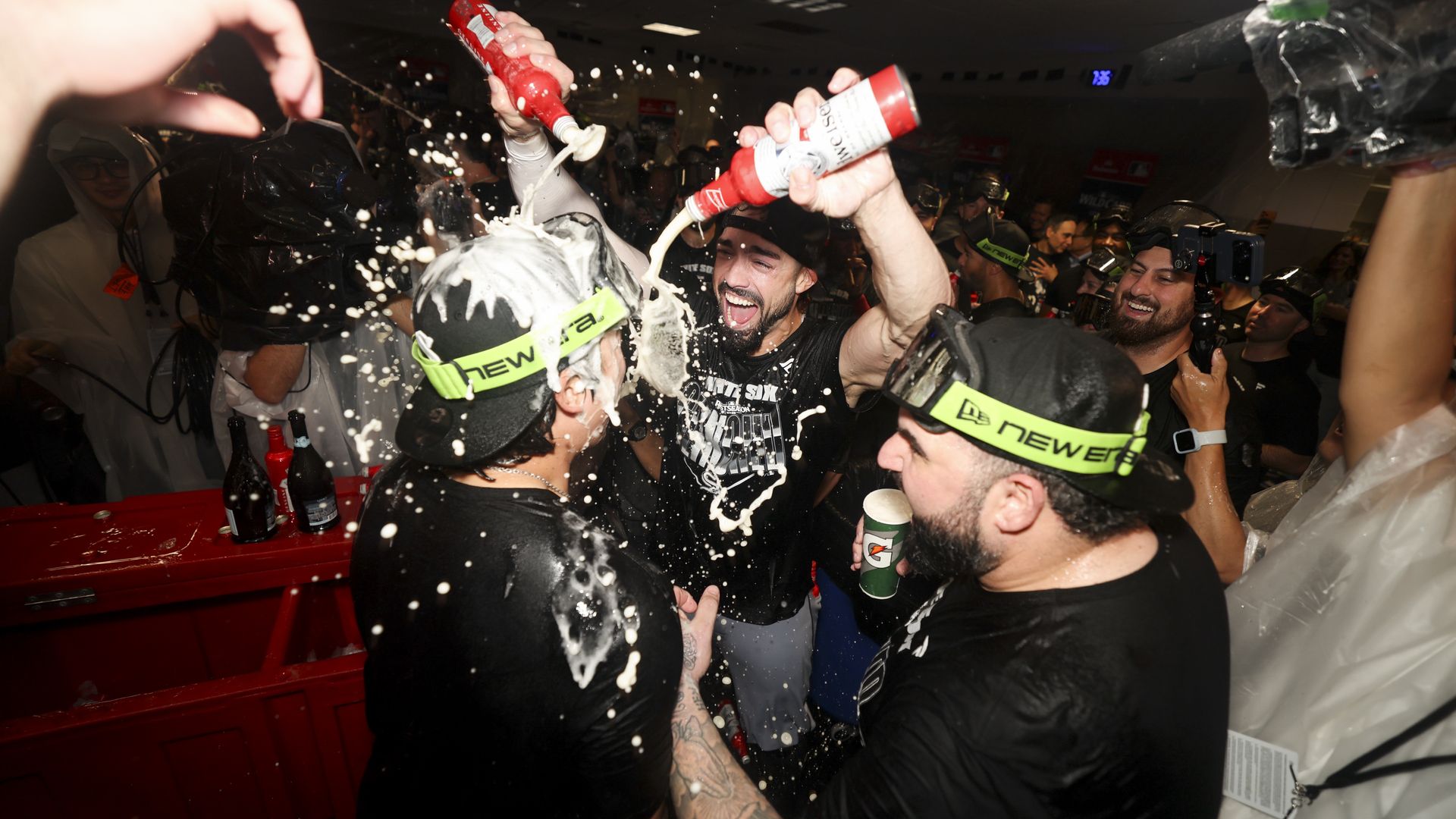 A crowded indoor party with people spraying foam from red bottles, laughing as foam splashes around. Participants wear neon headbands; festive chaos fills the dim room.