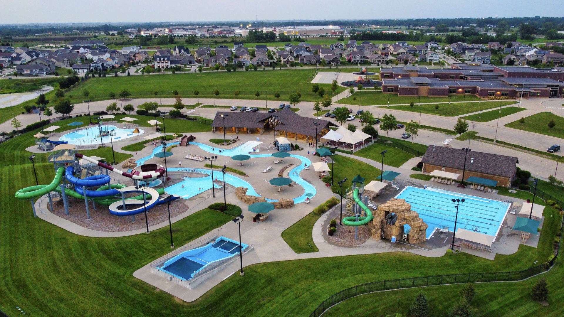 Aerial view of a community water park with winding blue and green slides, a lazy river, large pool, rock formations, shaded seating, and nearby suburban houses and parking lots.