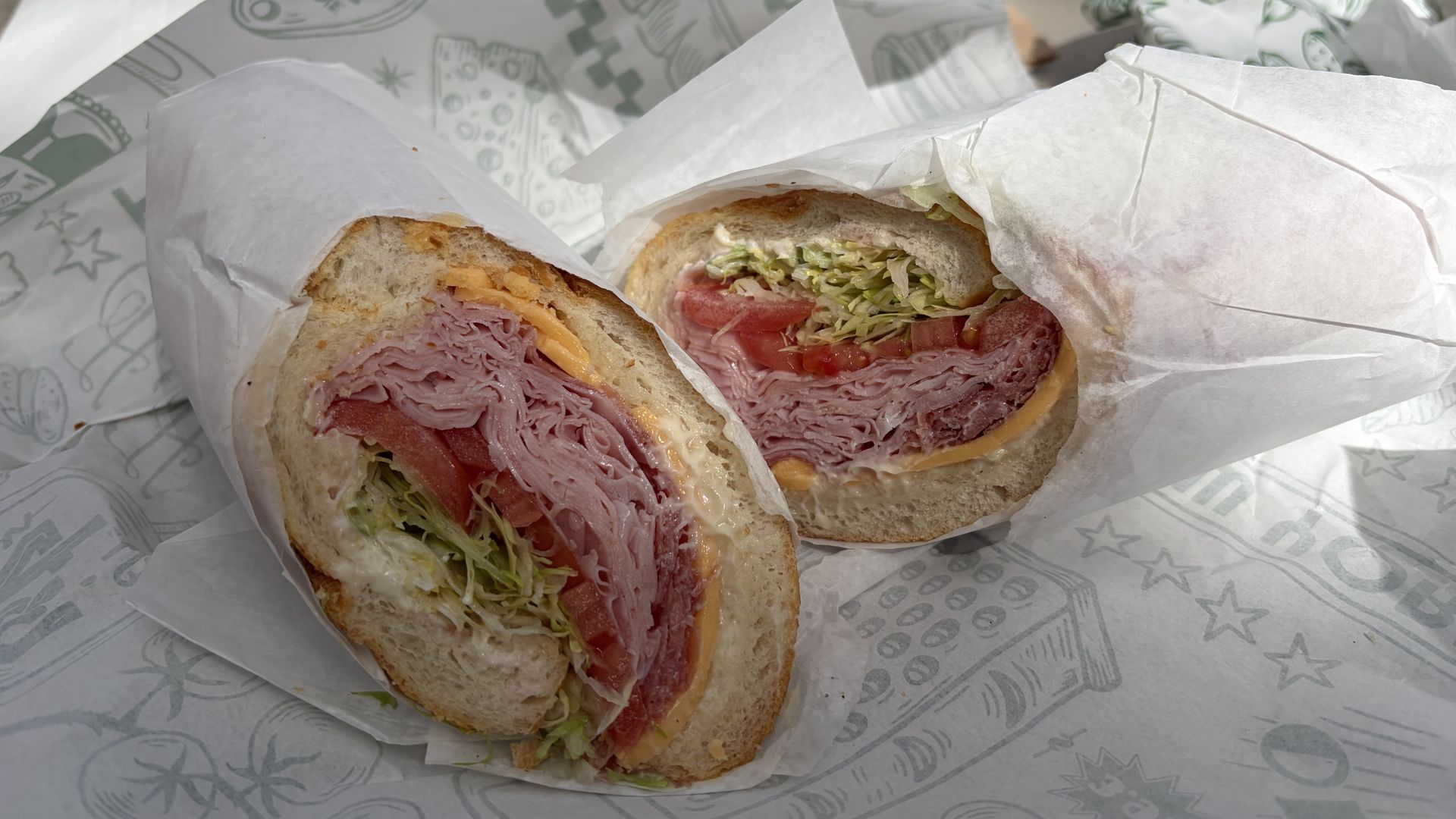 Deli sandwich halves wrapped in white paper showing layers of sliced ham, cheddar cheese, lettuce, tomato, and mayonnaise on a soft white sub roll with patterned deli paper underneath.