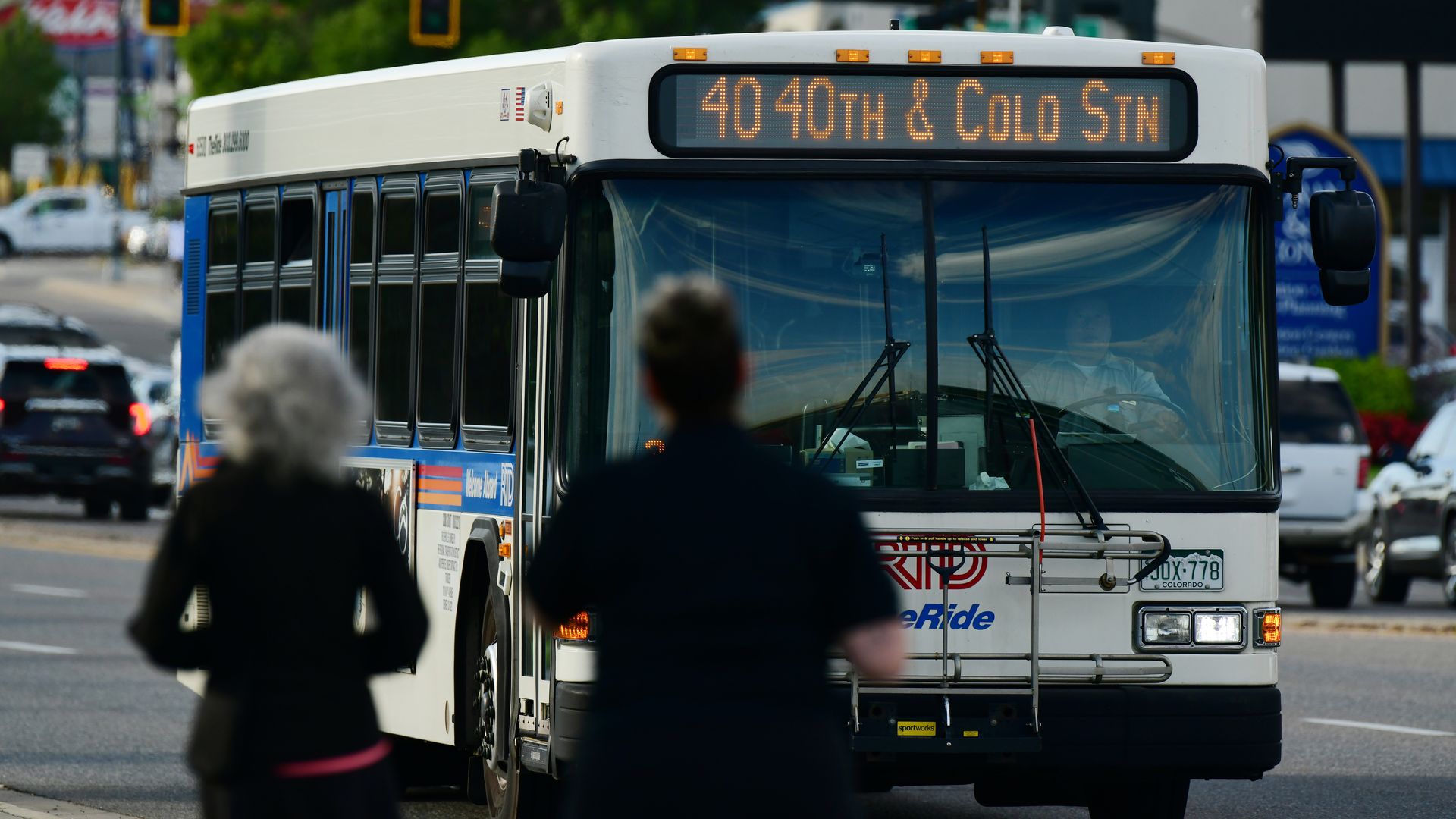 Front view of a white city bus on a busy street, with a digital route sign reading "40 40th & Collo Stn". Blurred pedestrians stand in the foreground as red traffic lights glow overhead.