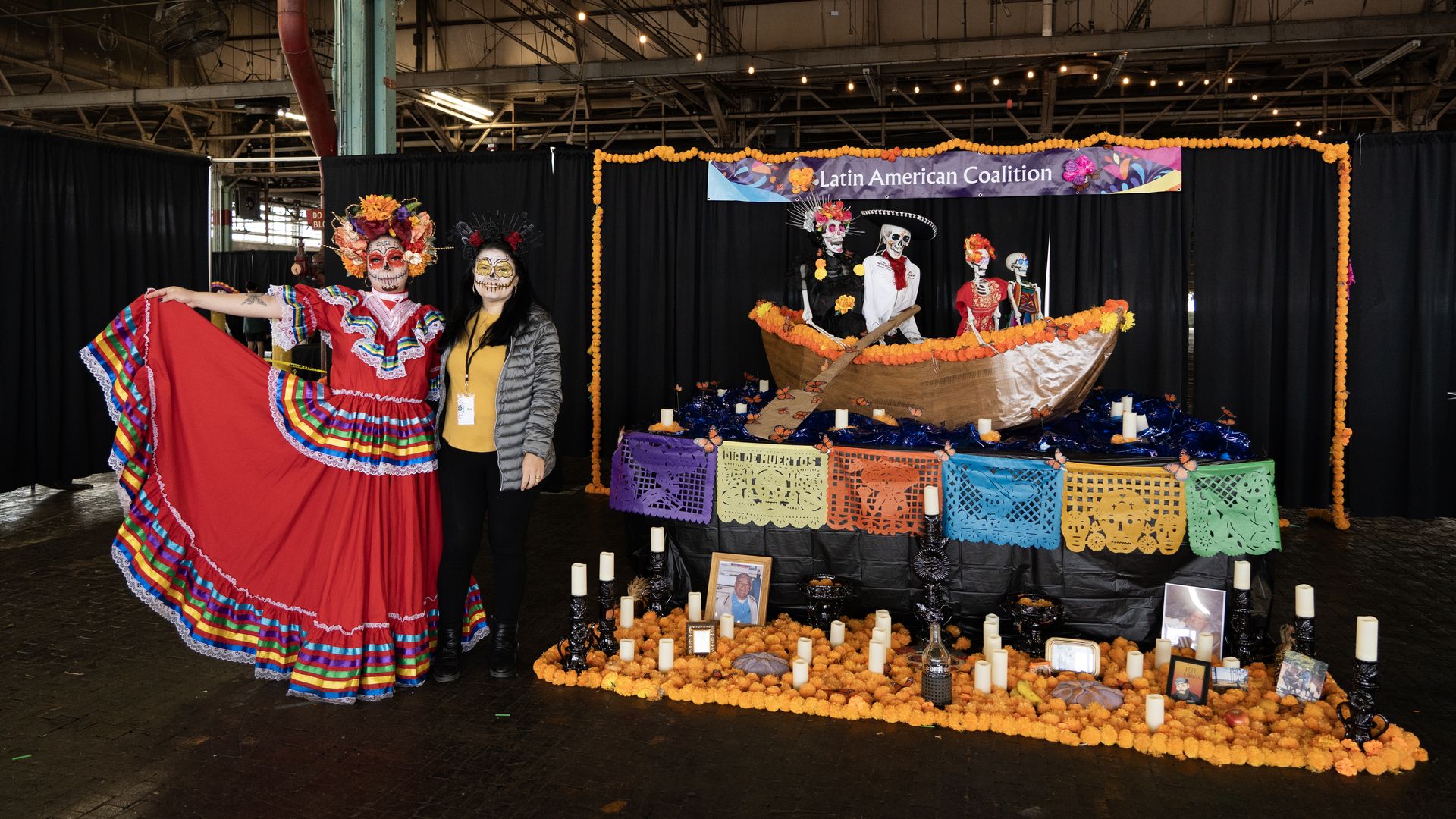 Two women with sugar skull face paint stand beside a colorful Day of the Dead altar decorated with orange marigolds, candles, papel picado, photos, and skeletons in a boat under a Latin American Coalition sign.