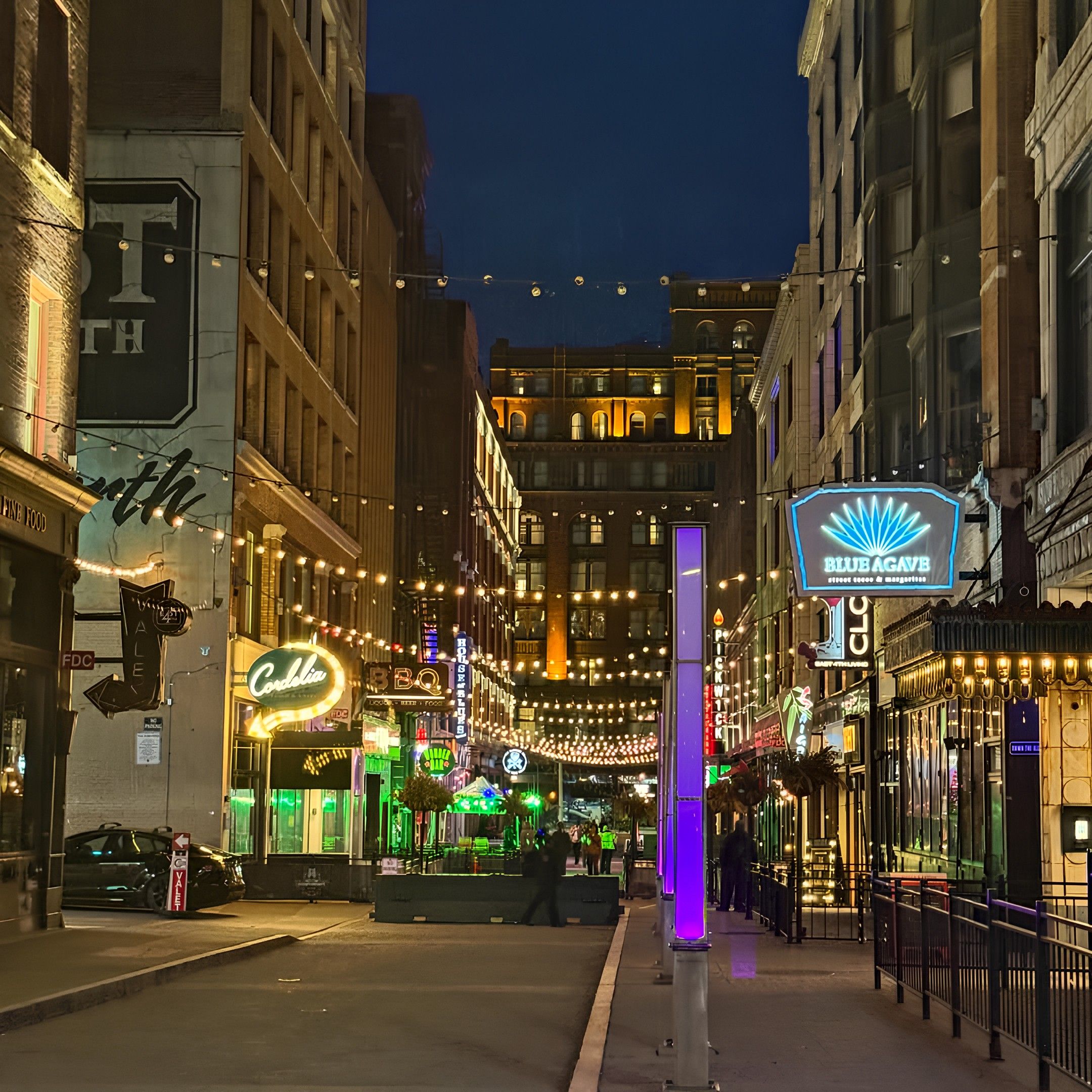 Night city street with string lights, a tall purple pillar in the center, and warm storefronts. Neon signs glow, including Blue Agave and BBQ, as pedestrians walk the sidewalk.