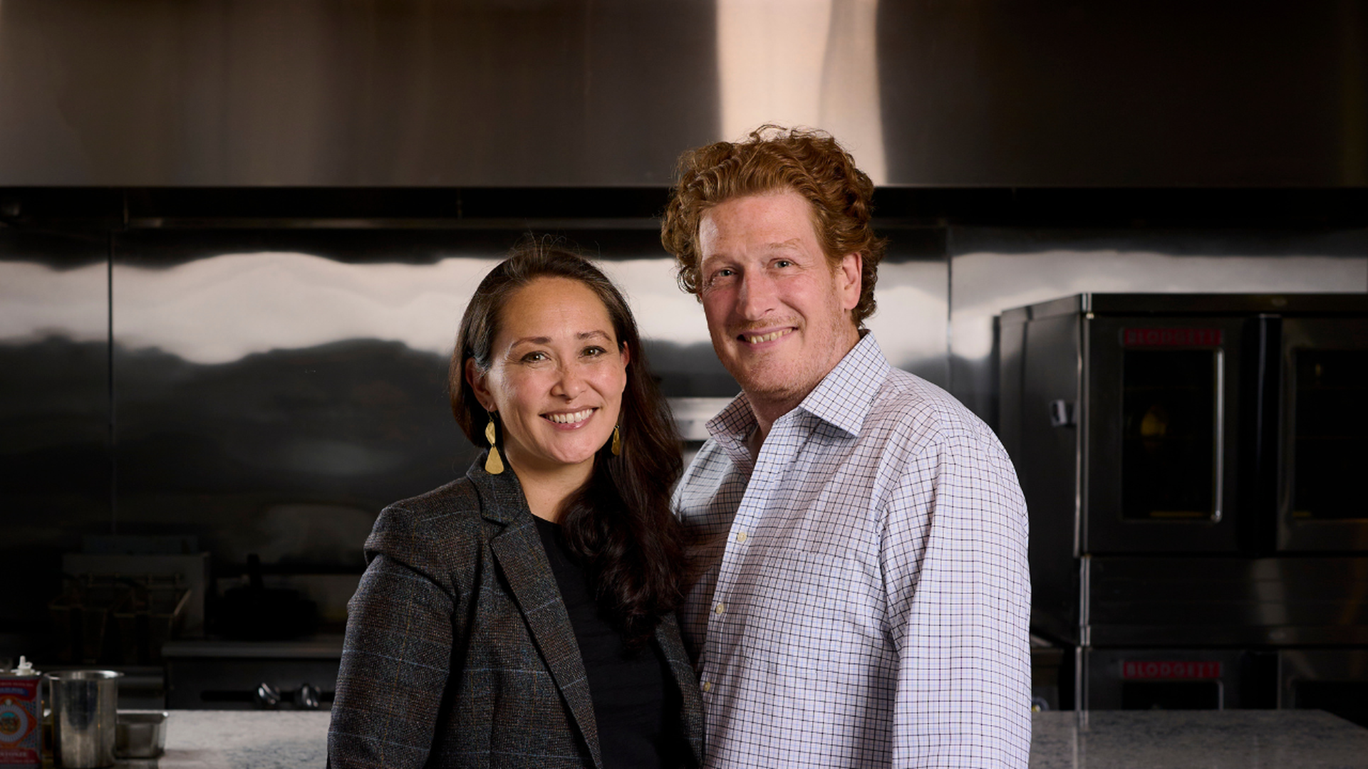 A smiling couple stands together in a stainless steel kitchen. The woman wears a dark blazer and gold earrings, the man a light checkered shirt. They pose closely and happily.
