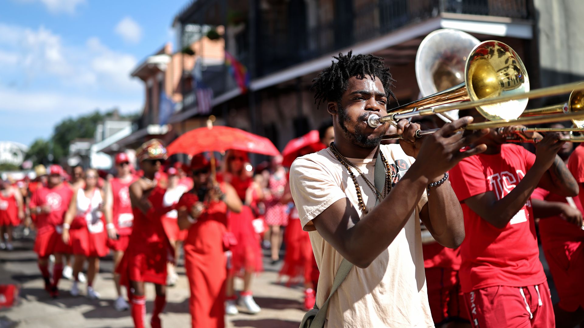 Man playing trombone leads a street parade with many people dressed in red, carrying red umbrellas, under a bright blue sky with some clouds.