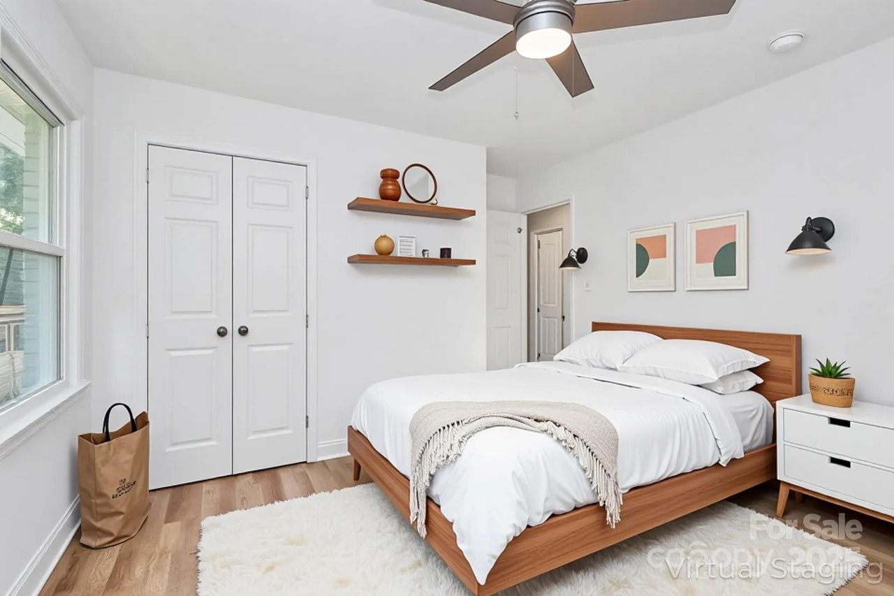 Bright bedroom with white walls, wood bed frame with white bedding, a beige throw blanket, two abstract art pieces above the bed, wooden shelves with decor, and a white rug on wood floor.