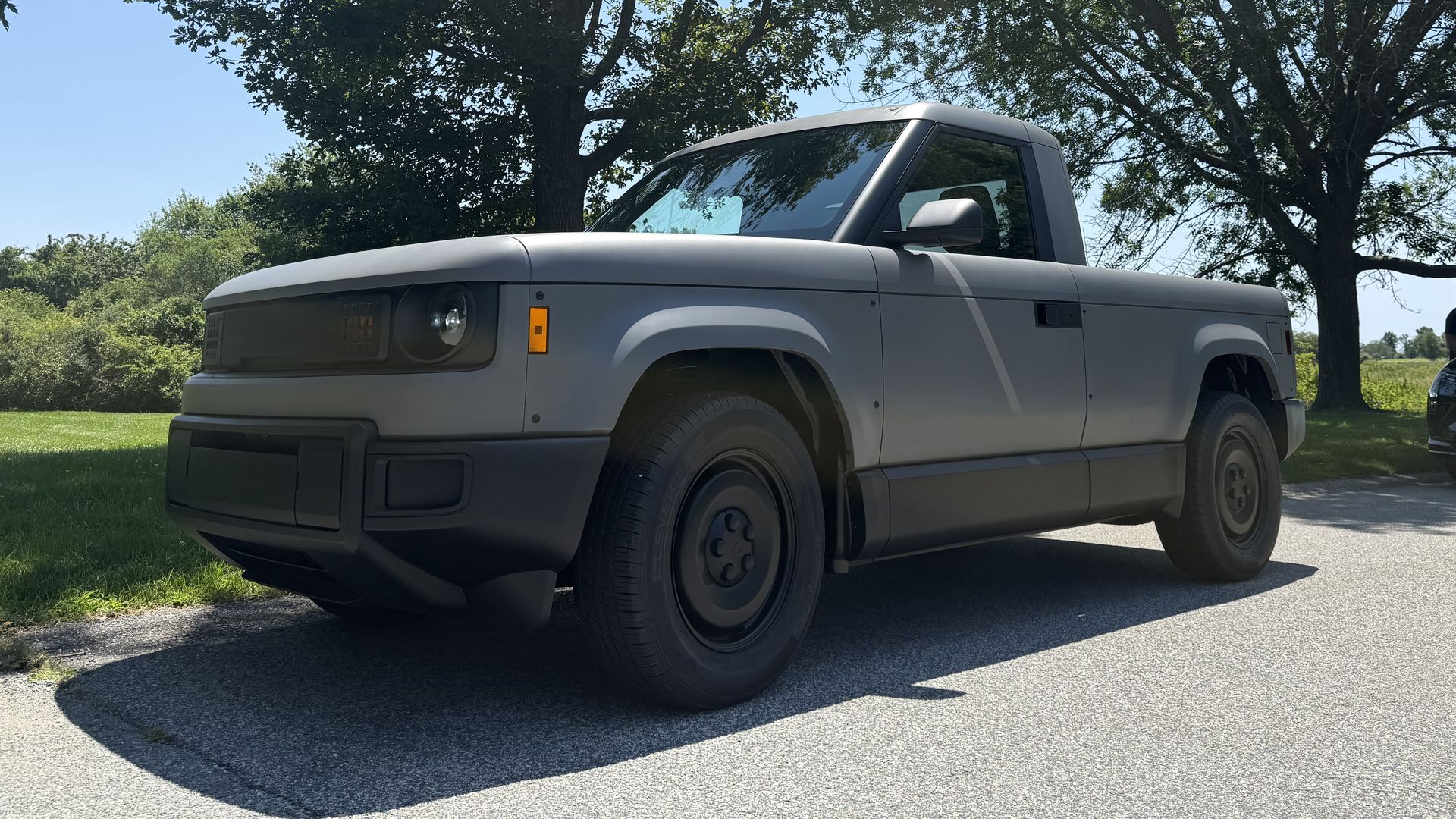 A gray Slate Auto electric pickup prototype is parked on the side of a road, by leafy trees, an open grassy field, and a clear blue sky.
