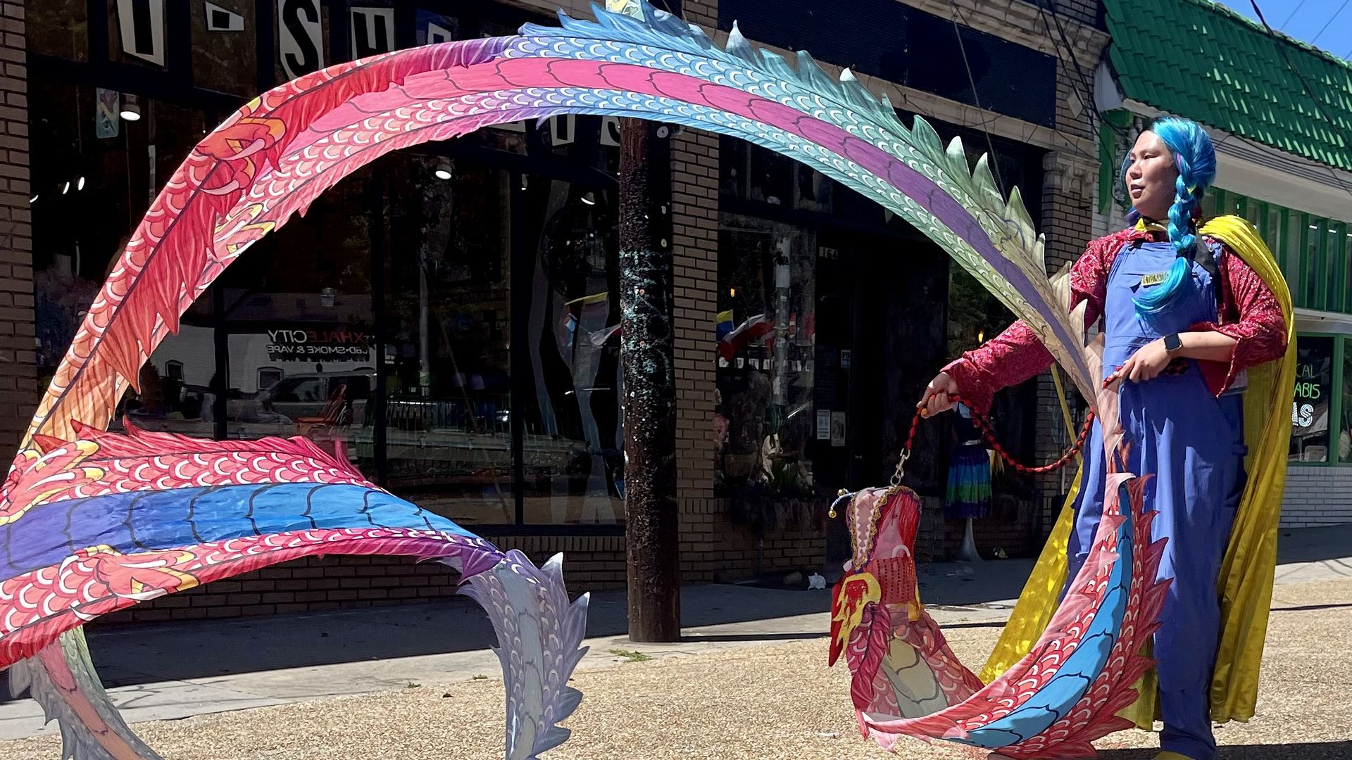 A performer wearing a blue wig and blue outfit, guiding a long multicolored dragon with red, pink, blue, and green scales; brick storefronts and a green roof in the background.