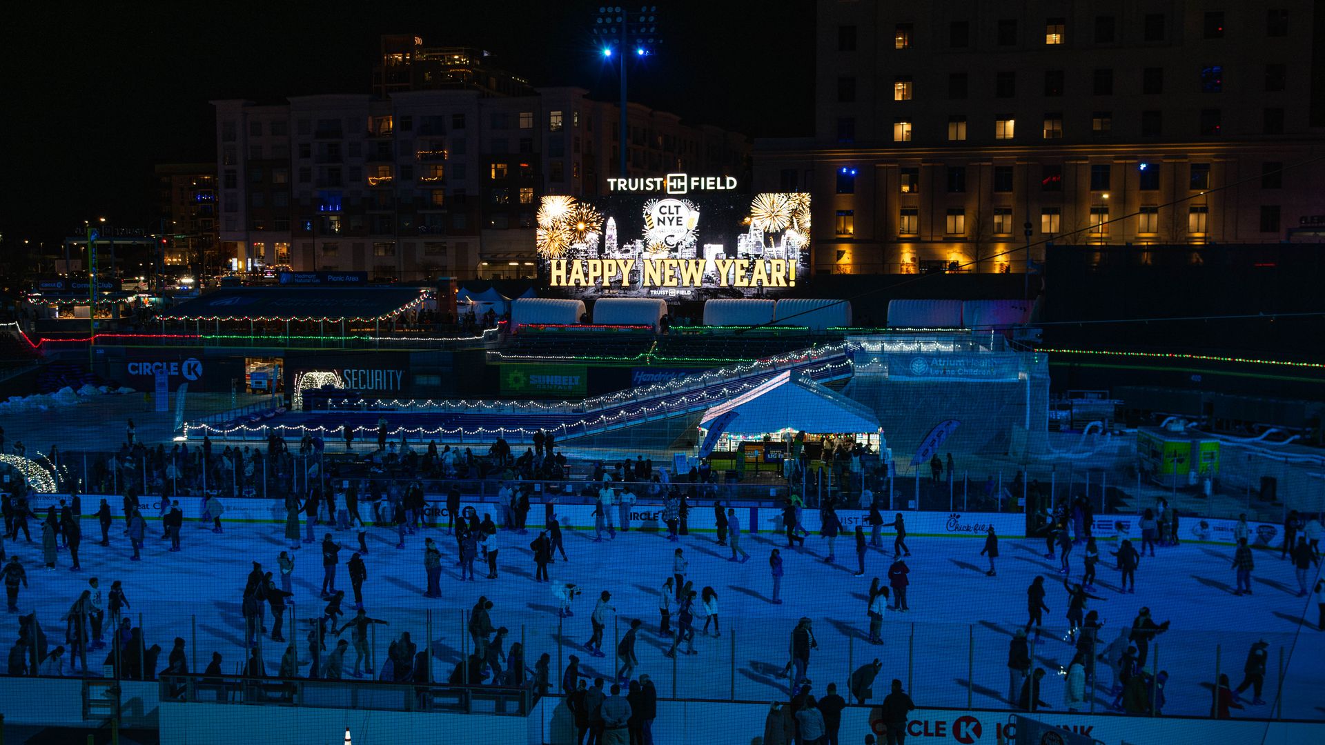 Nighttime ice skating at Truist Field with many people on the ice, holiday lights, and a large illuminated sign saying "HAPPY NEW YEAR!" and "CLT NYE" with fireworks graphics.