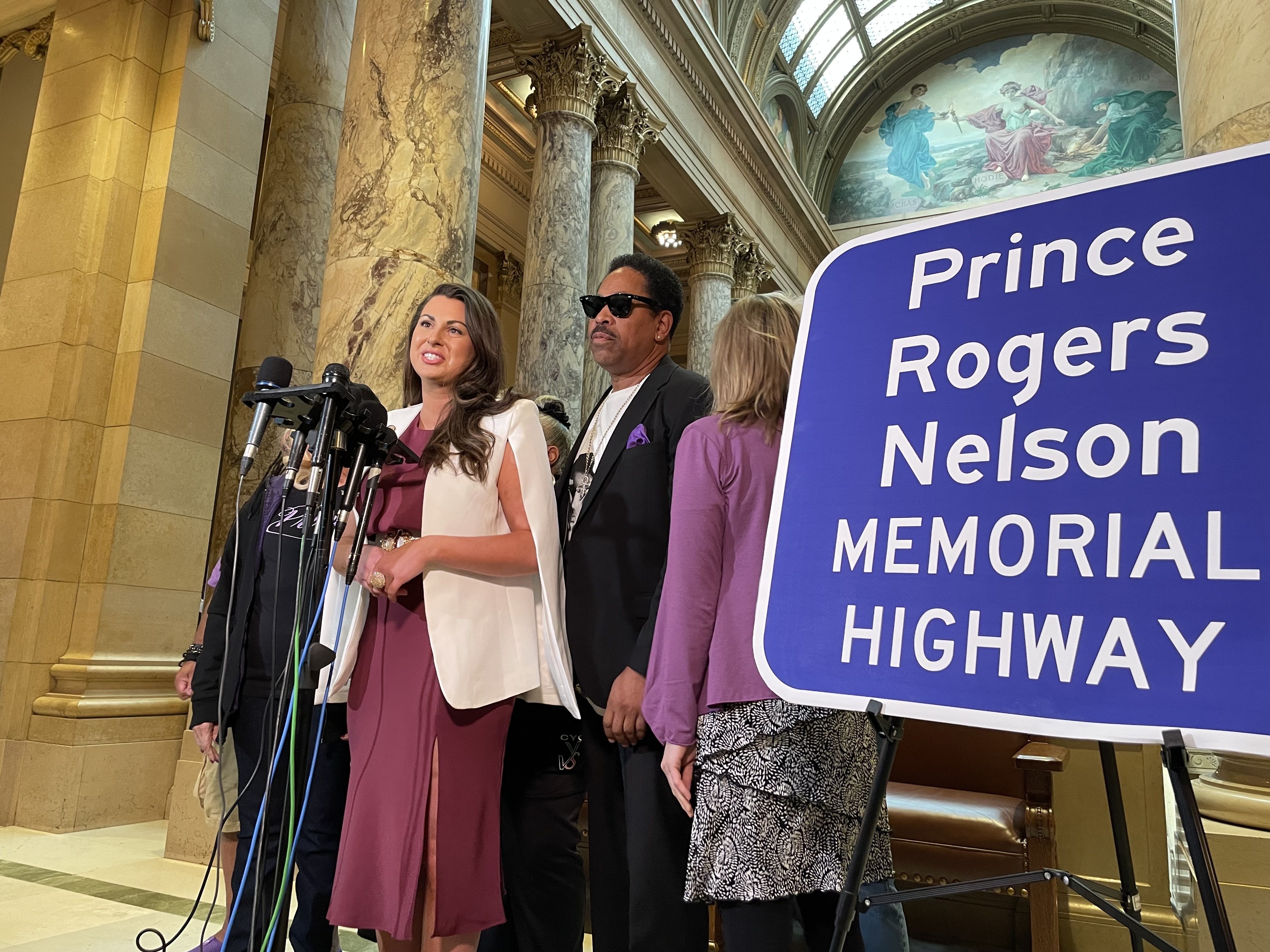 state senator in a urple dress and white jacket stands at a microphone in front of a prince highway sign