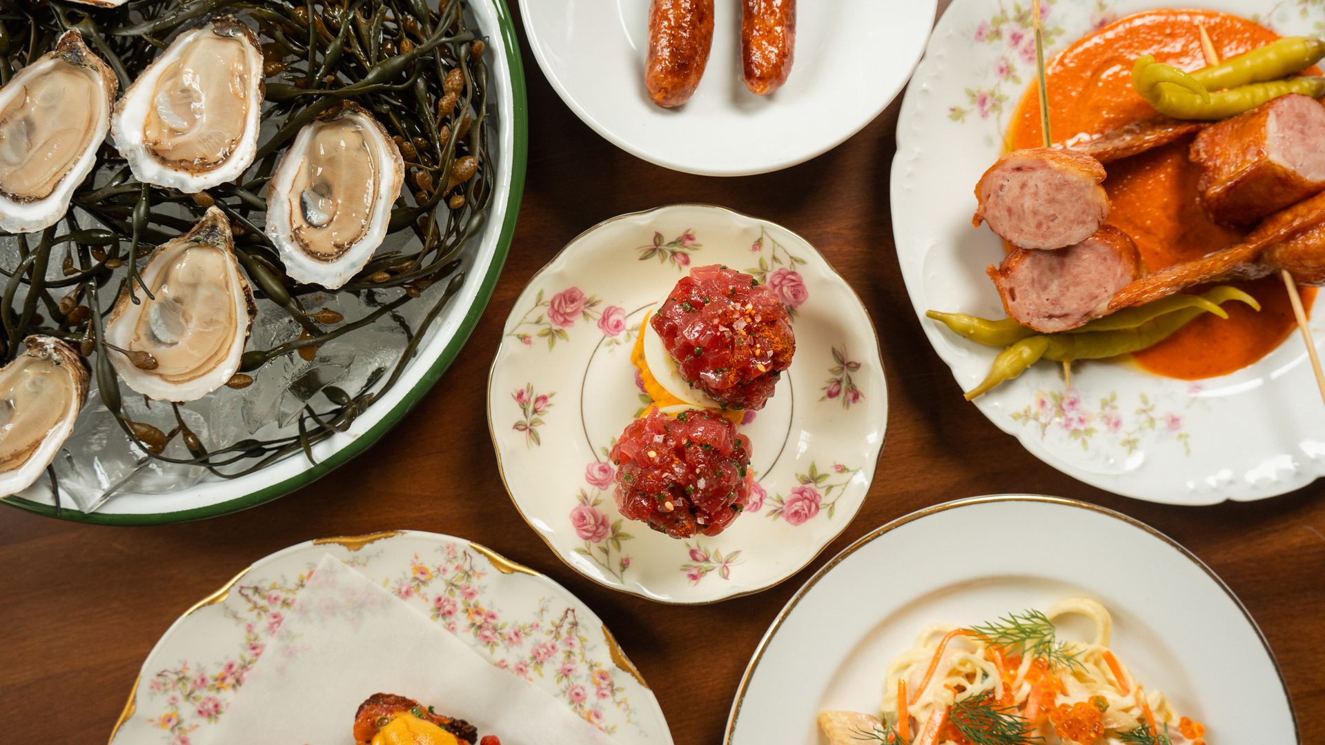 Assorted dishes: oysters on ice with seaweed, two sausages on a white plate, tuna tartare on a floral plate, and sliced sausage with orange sauce and asparagus on another floral plate.