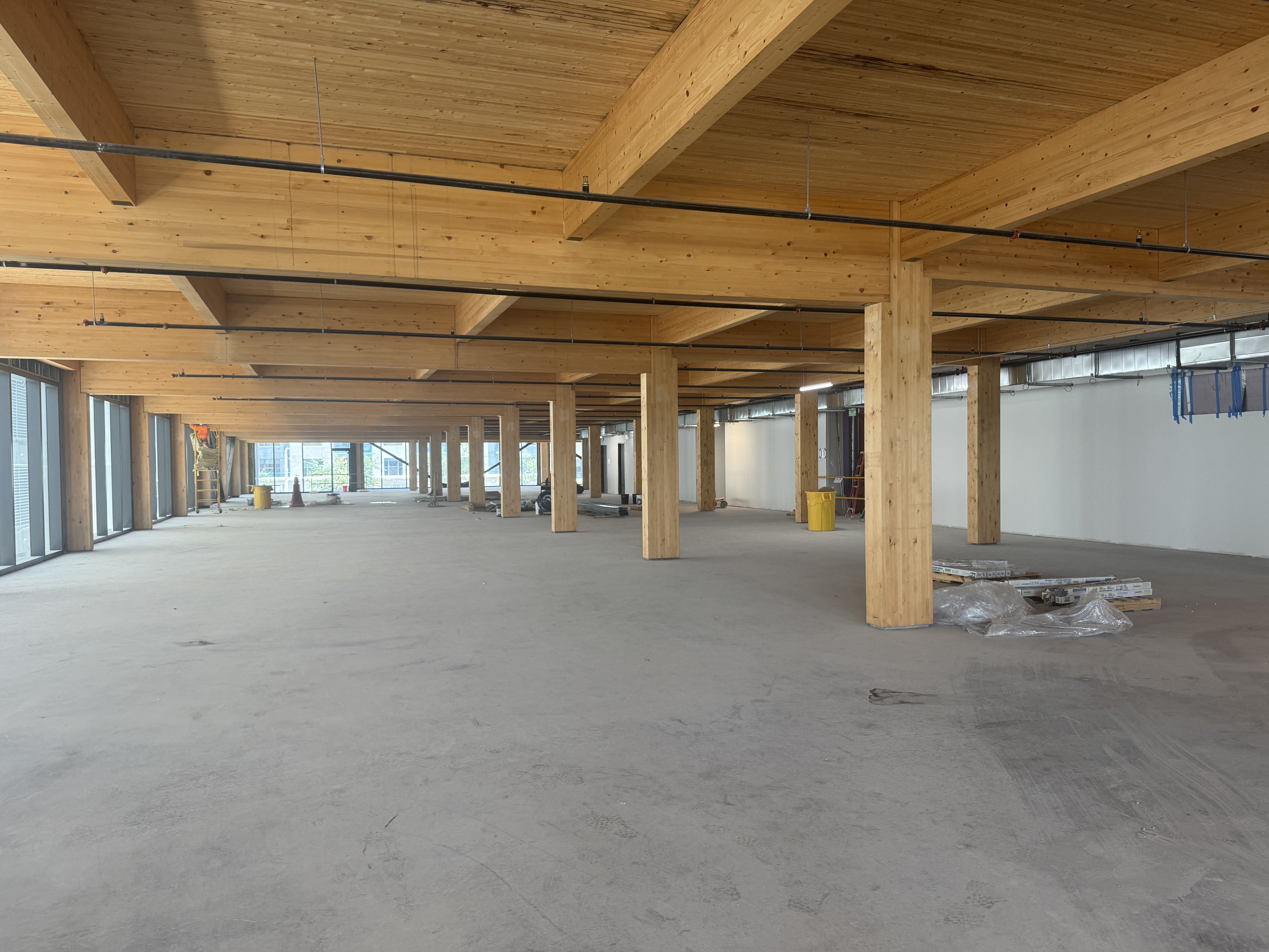 Interior of a large construction site with exposed wooden beams and columns, black overhead pipes, and a concrete floor. Windows line the left, with yellow bins and pallets nearby.