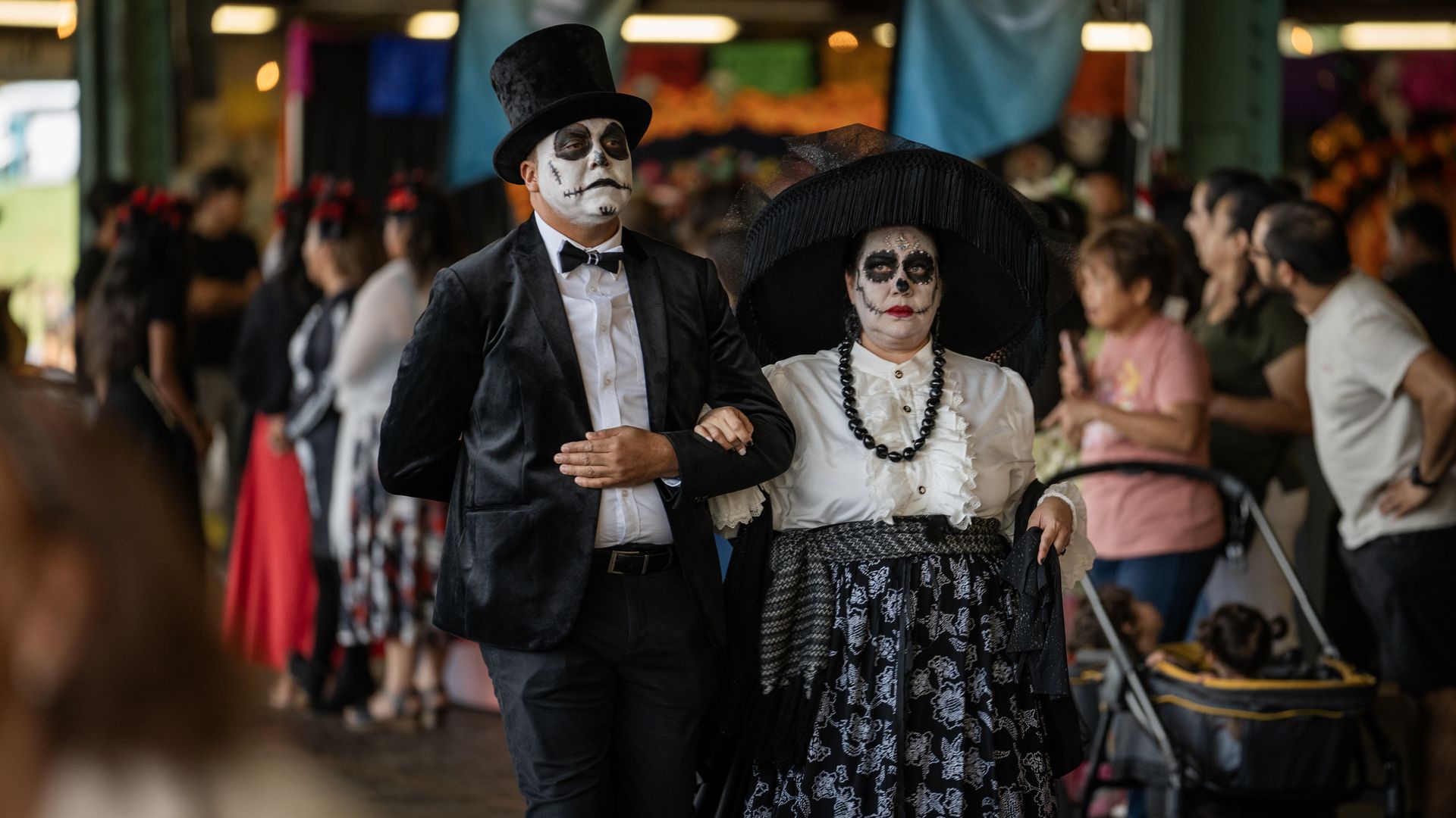 Couple in Day of the Dead costumes walk arm-in-arm through a crowded market: man in a black suit and top hat with skull makeup, woman in a large black hat and white blouse with lace skirt. Halloween things to do in Charlotte this weekend 2026