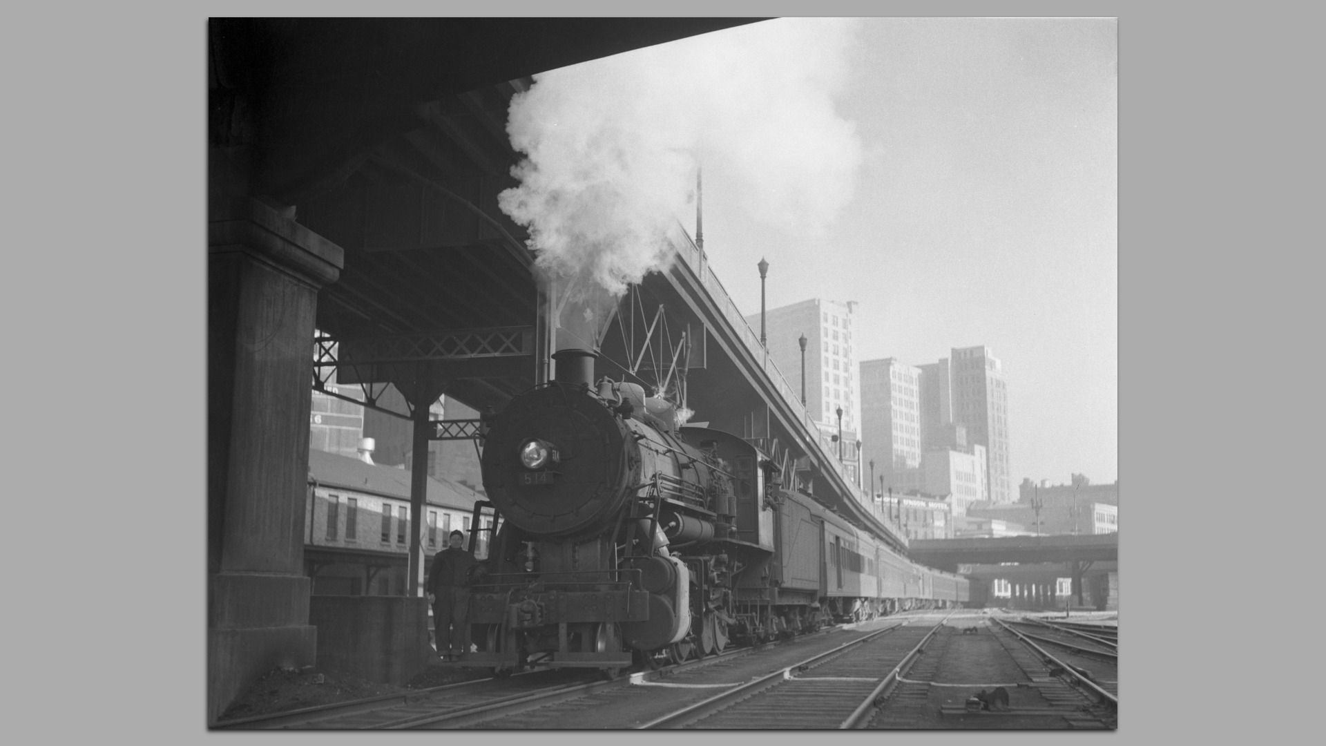 A black and white photo of a train with exhaust or steam billowing from the locomotive travels under a viaduct