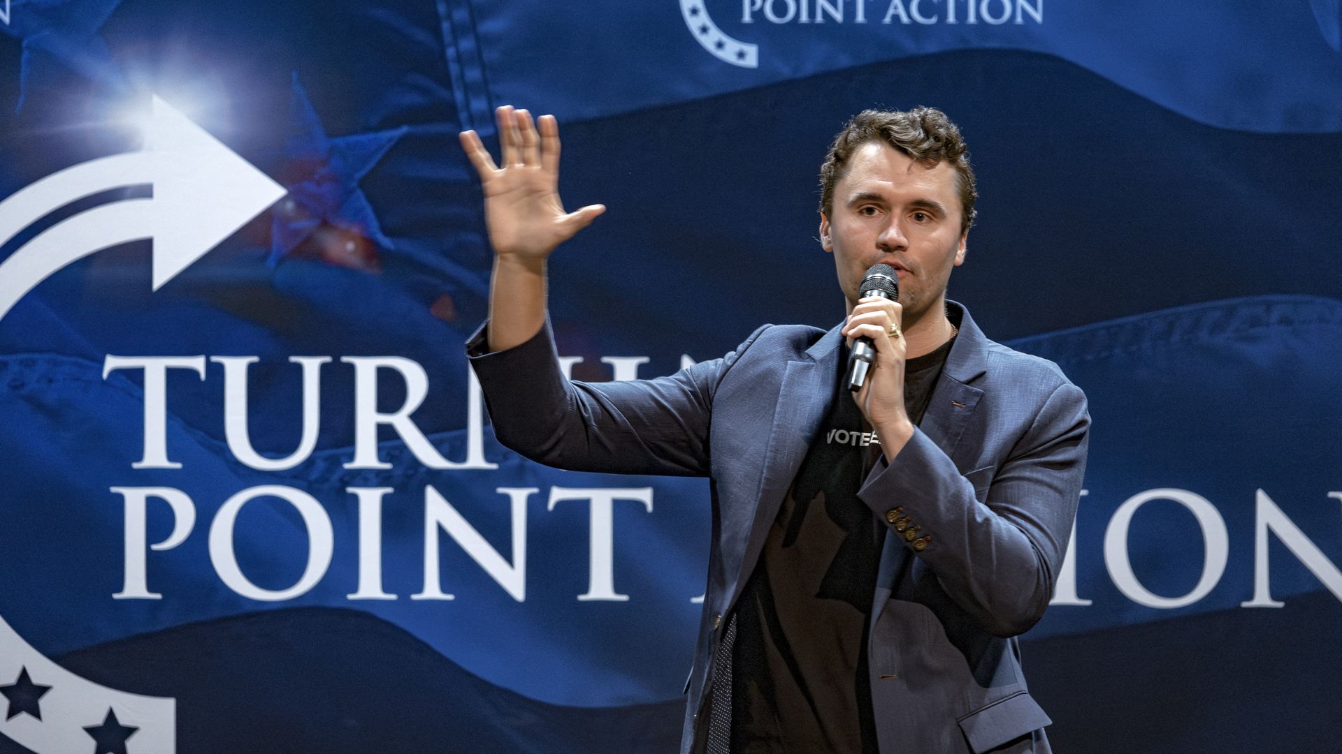 A man raises his hand and holds a microphone in front of a backdrop that says "Turning Point Action."