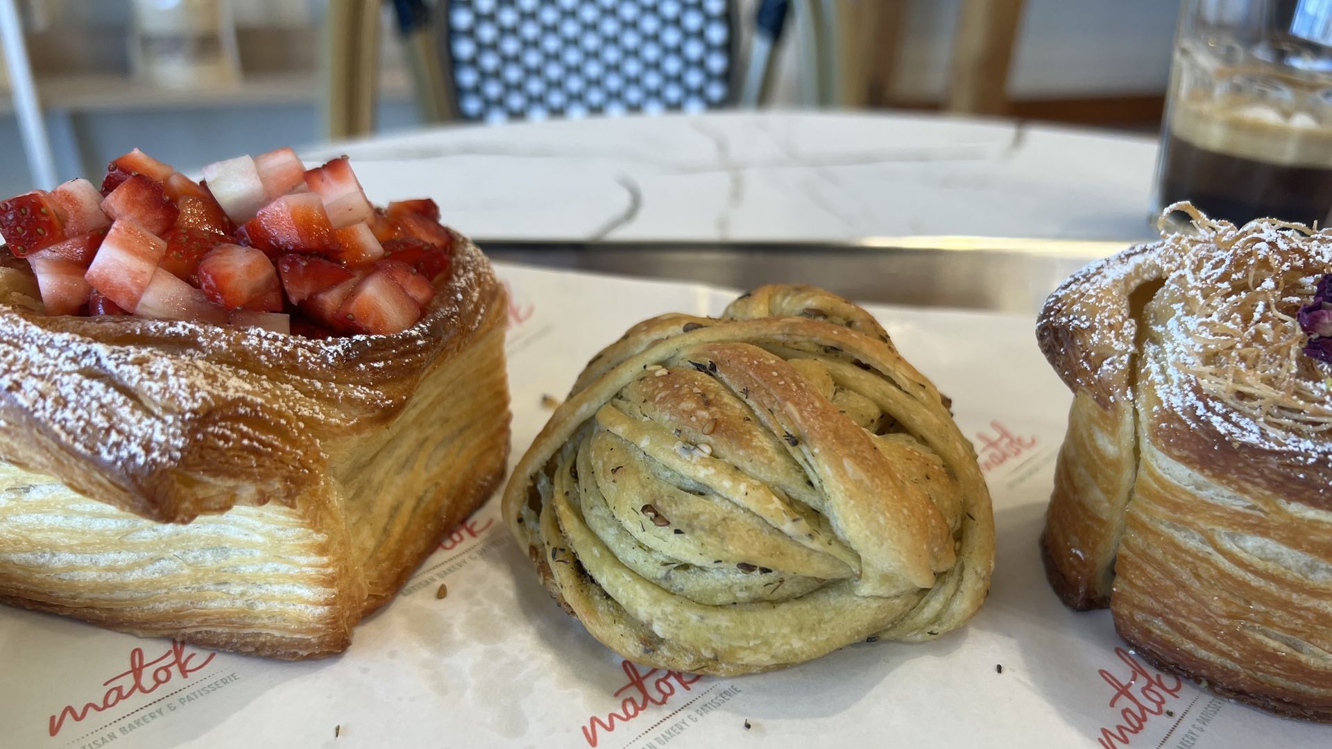 Three pastries on white paper with red "matok" logo: left square puff topped with diced strawberries, middle twisted seeded bread, right square puff with shredded topping and powdered sugar.