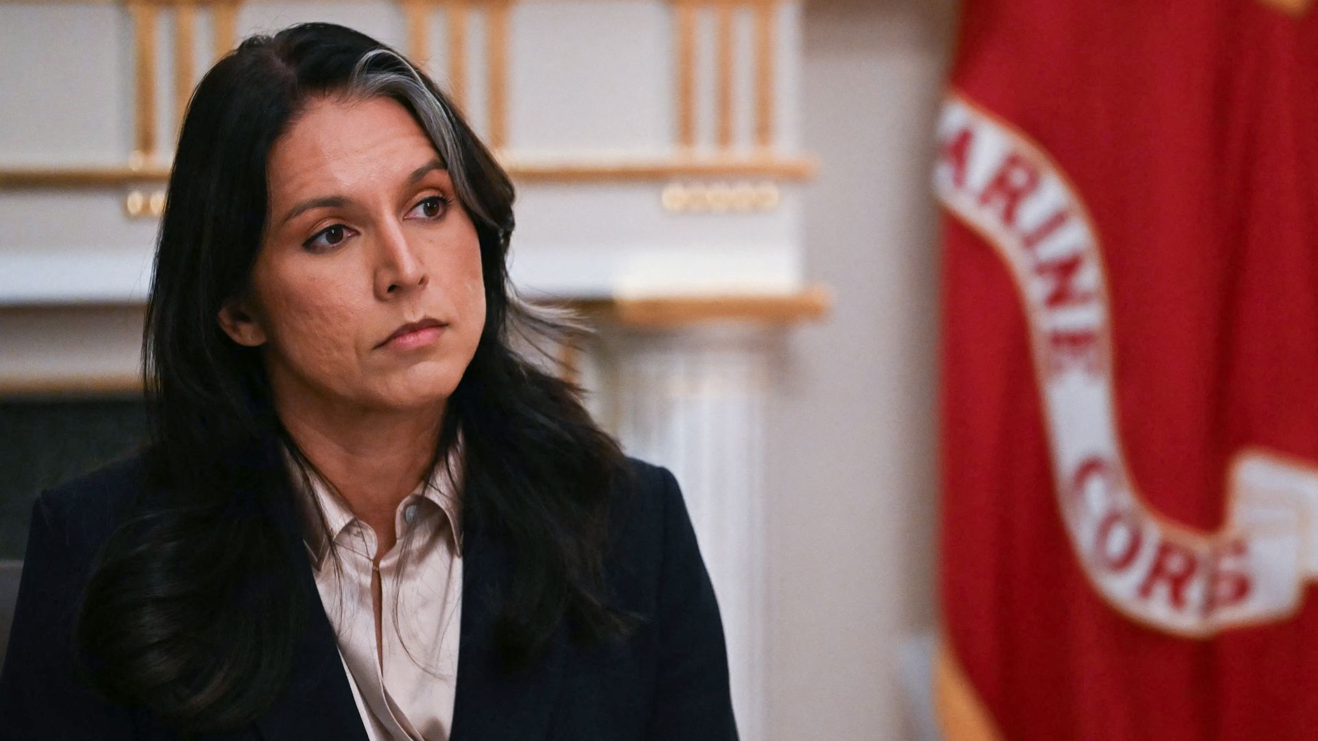 Director of National Intelligence Tulsi Gabbard, with long dark hair and a black blazer with gold buttons sits indoors, looking to her left thoughtfully, with a red Marine Corps flag partially visible in the background.