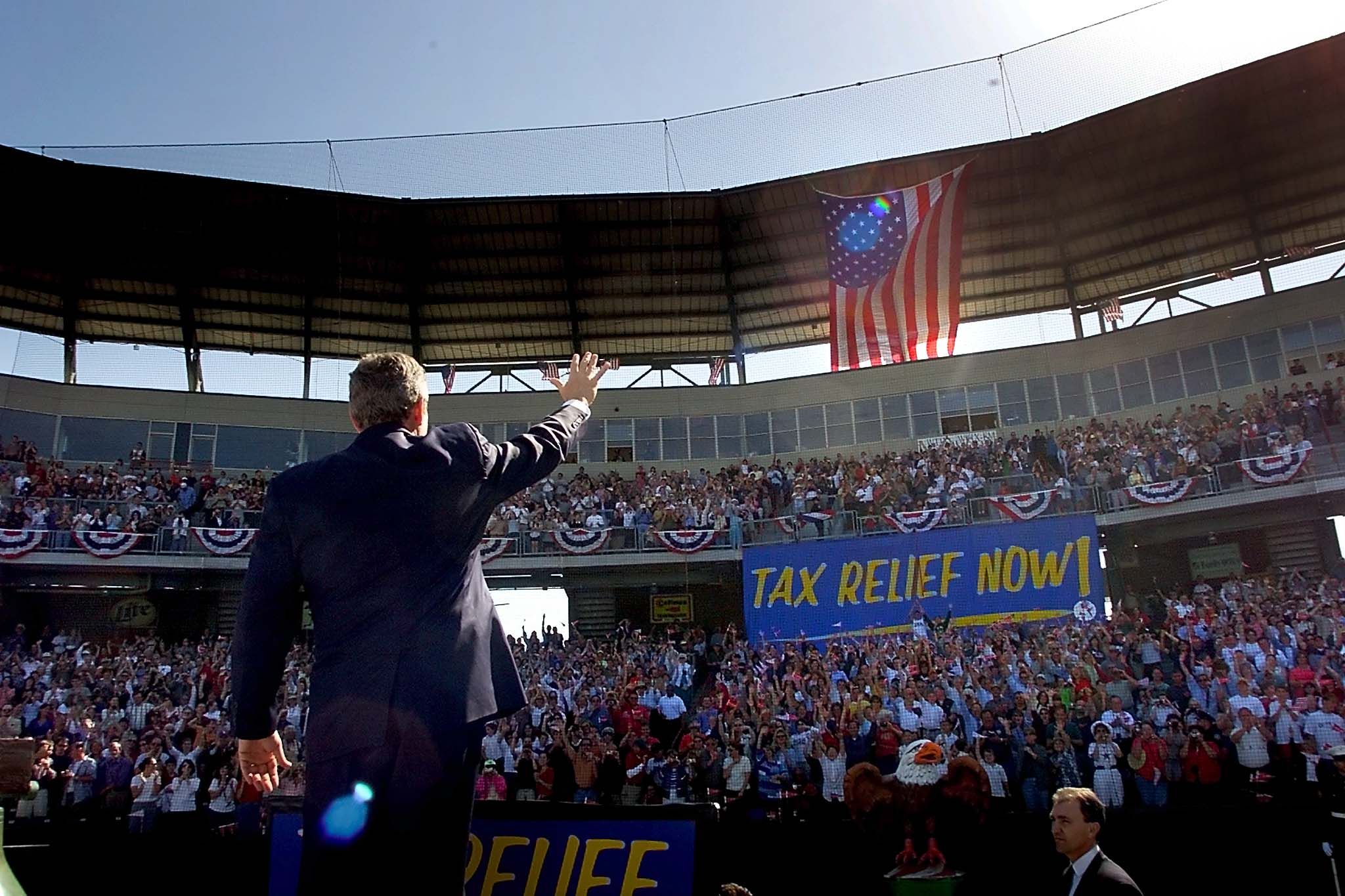 President George Bush is seen from behind as he waves to a crowd of people in a baseball stadium. A large sign hangs from the rafters and says "Tax Relief Now!"