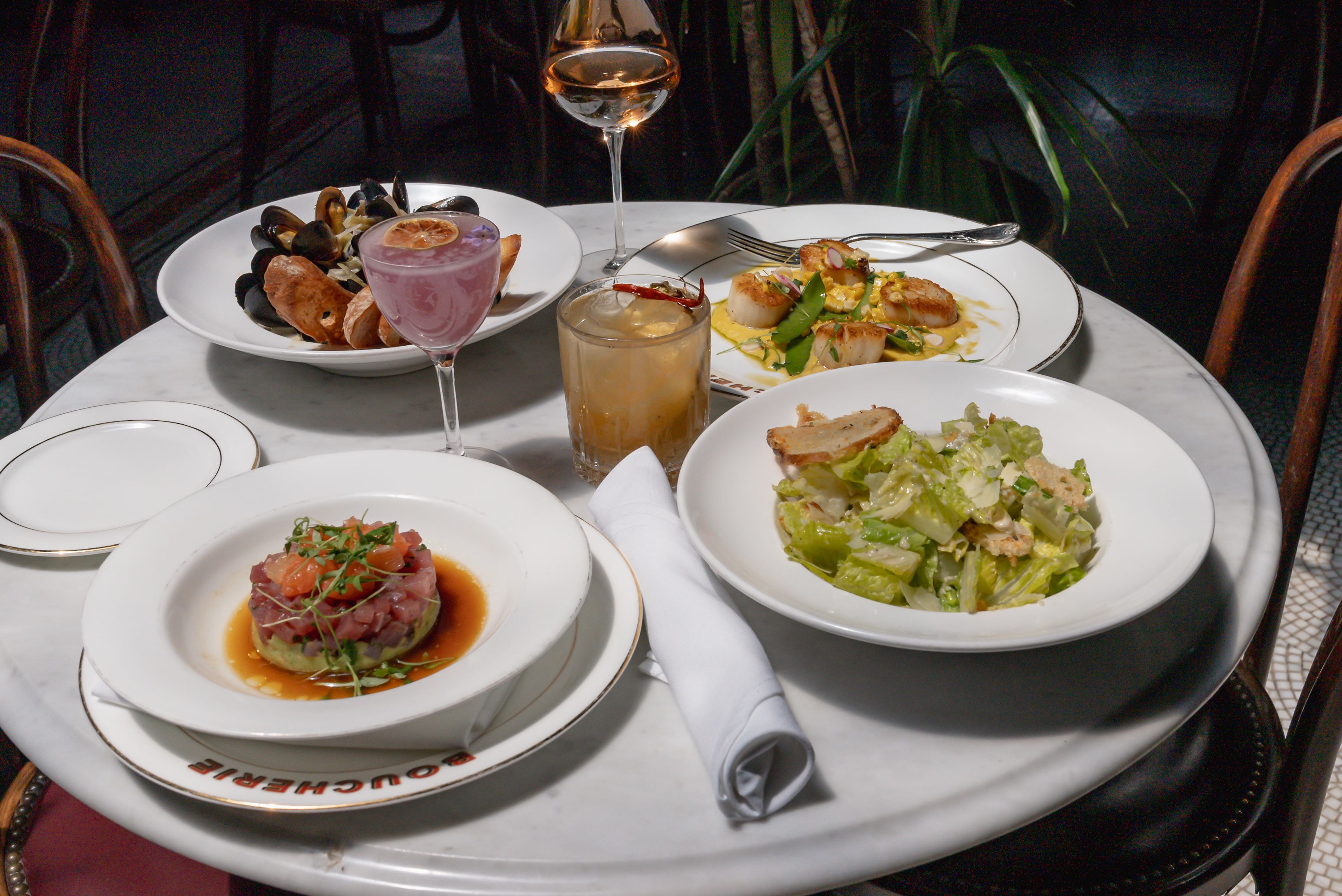 Four while bowls of food on a round table-clothed table with a cocktail, low ball glass cocktail and glass of wine.