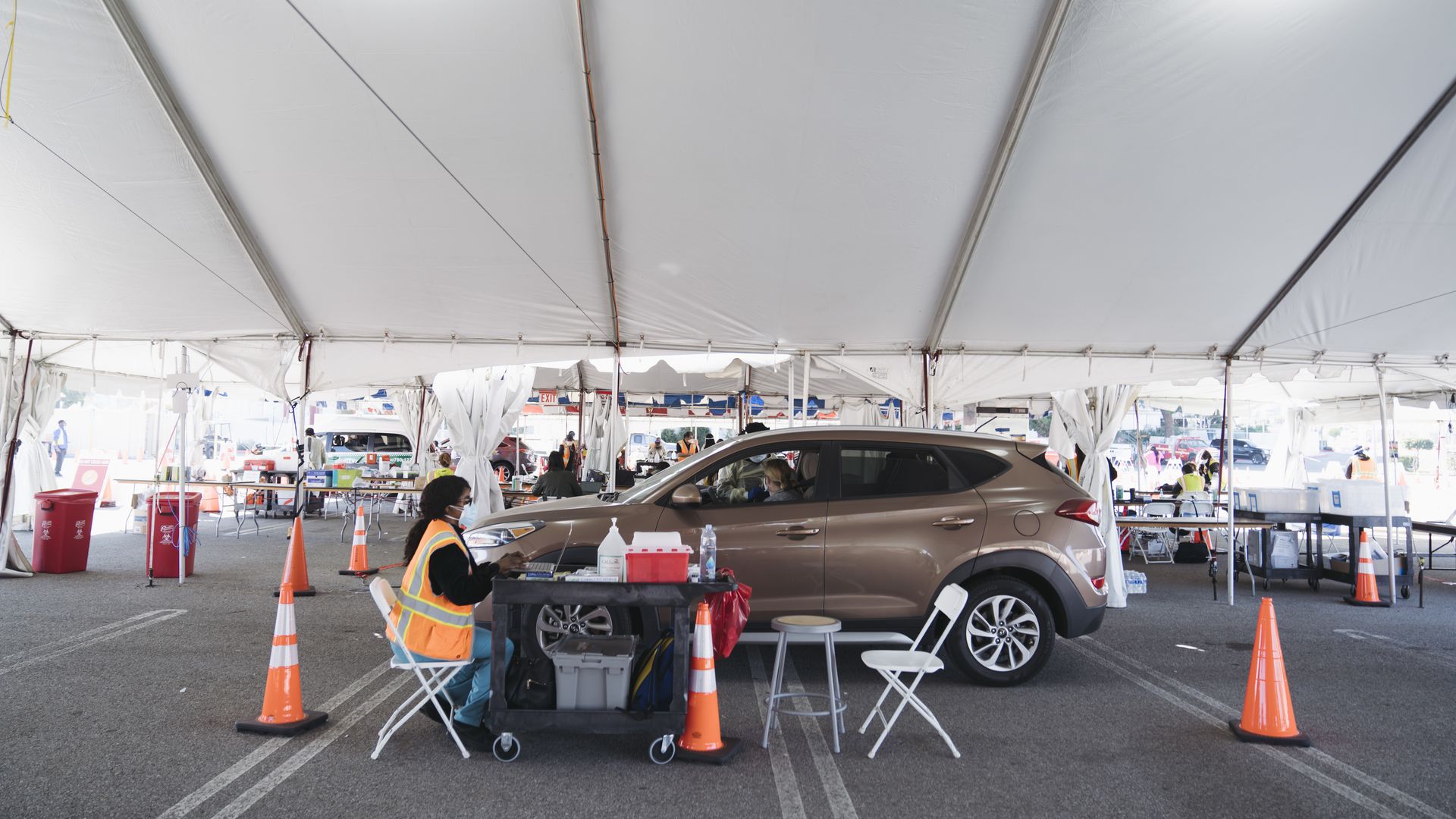 A car is parked next to a woman sitting in an orange vest at a table 