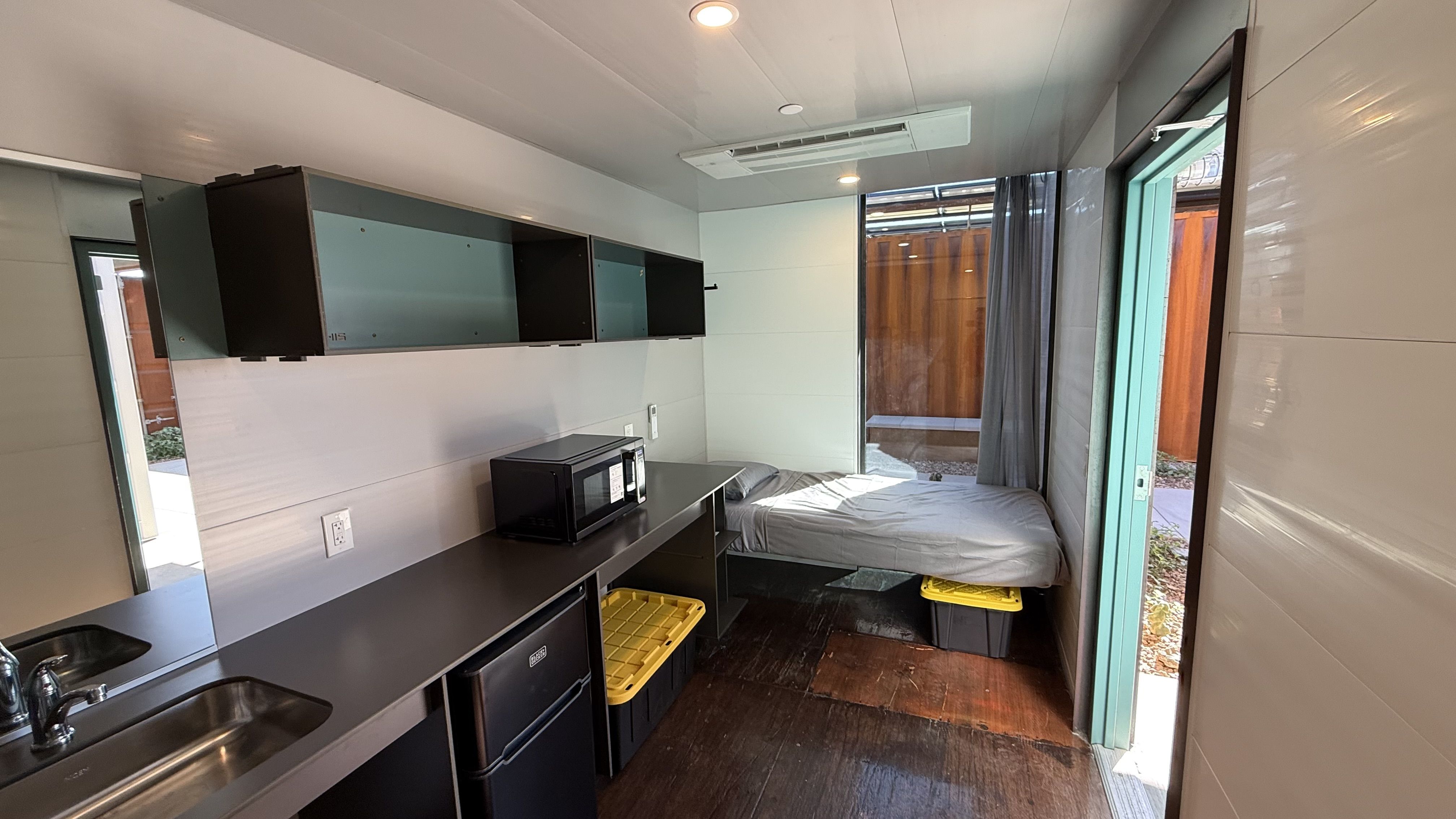 Small dormitory-style room with a sink and microwave on a metal counter, and teal/black wall cabinets above and a small refrigerator underneath. At the back there's a bed with grey sheets. 