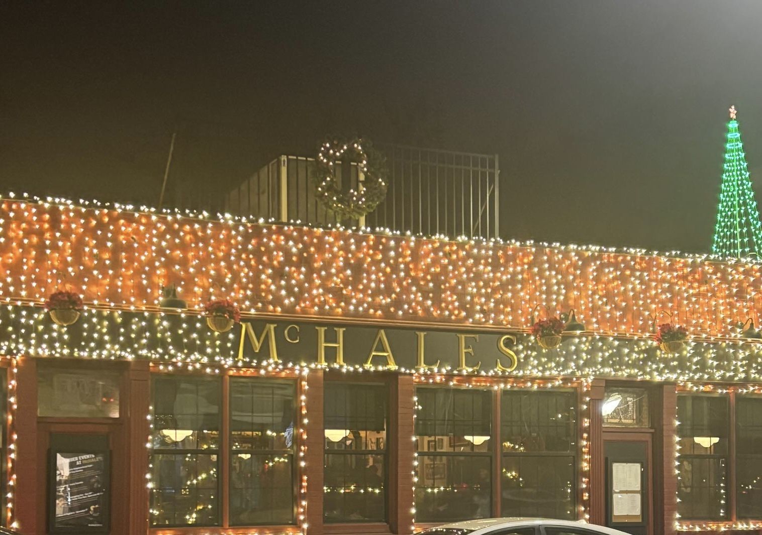 Building with a sign reading McHALES decorated with white and orange Christmas lights, a lit green Christmas tree decoration on the roof, and a wreath above the sign at night.