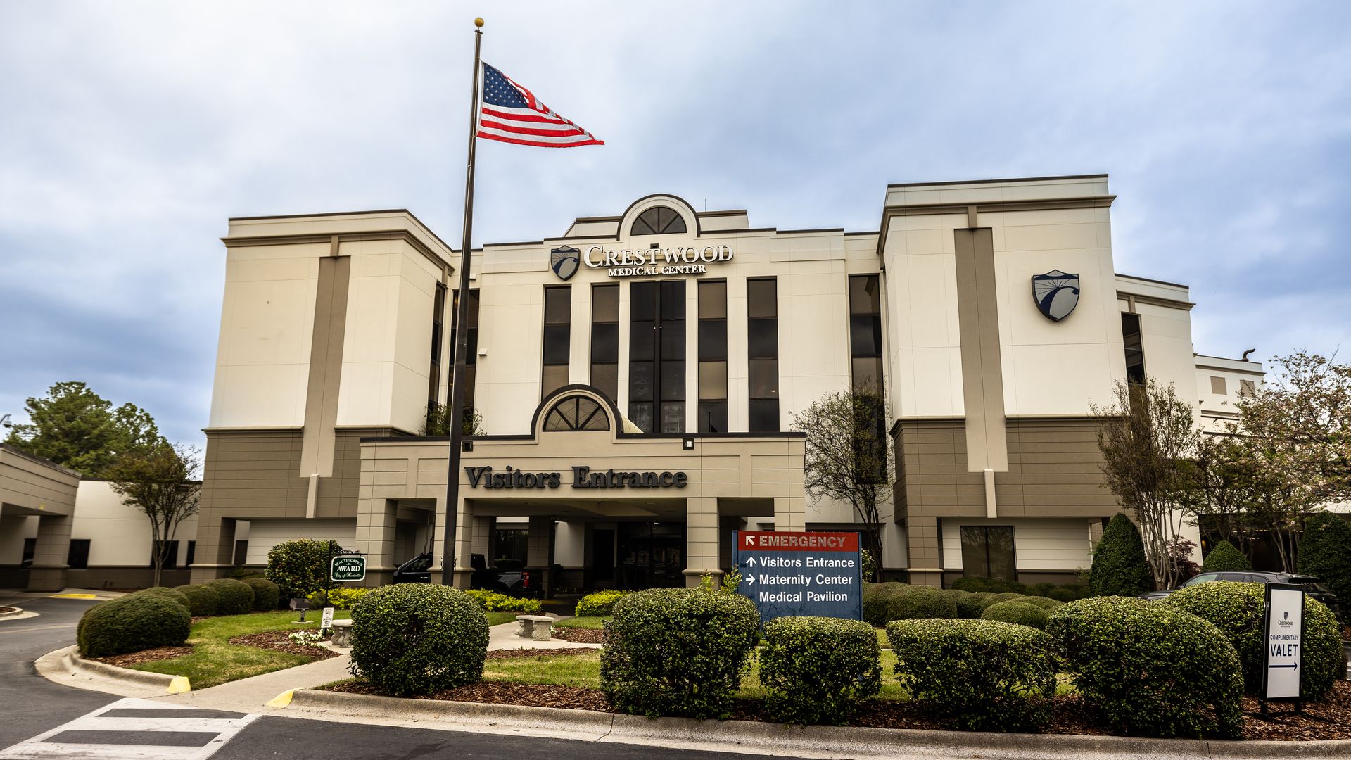 Exterior of Crestwood Medical Center, showing the Visitors Entrance under a cream-gray facade; an American flag on a tall pole, landscaped shrubs, and a blue shield logo on the building.