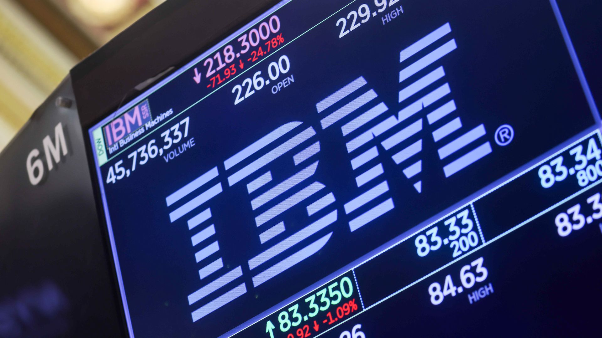 Close-up of a stock market monitor: blue screen with white numbers, red down values and a large downward arrow; IBM logo in the lower-left and diagonal white bar graphics.