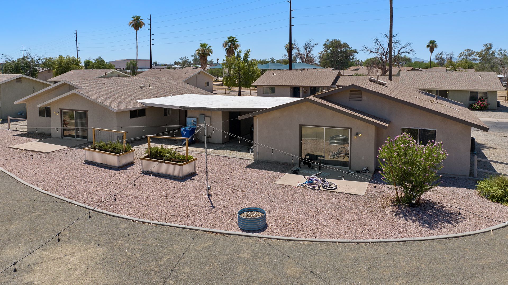 Single-story beige houses with brown roofs under clear blue sky, gravel yards, string lights, two bicycles, planters with greenery, and palm trees in the background.