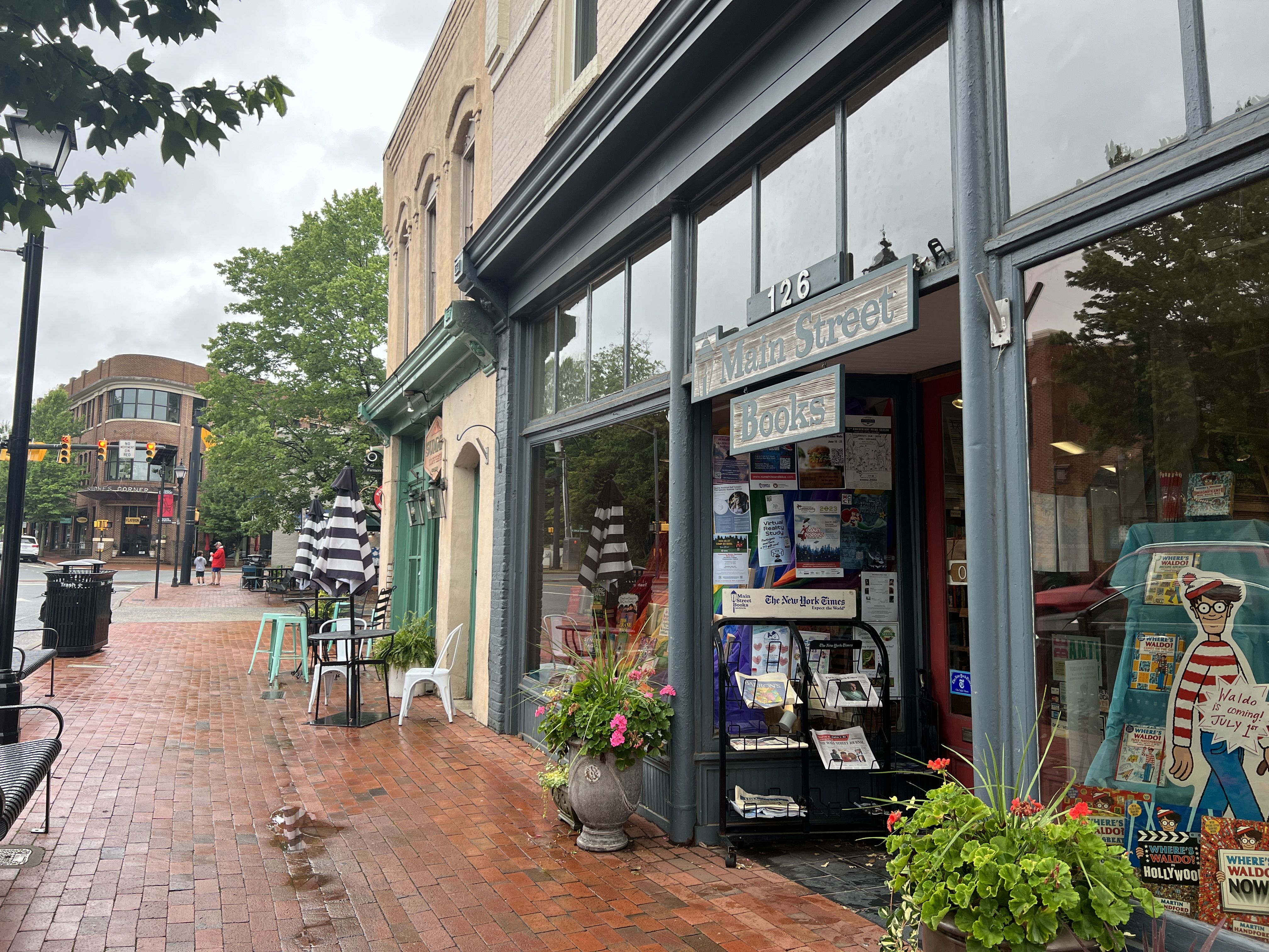 Brick sidewalk street view showing Main Street Books storefront with display racks, plants, and a Waldo poster, striped umbrellas, chairs, and buildings under cloudy sky.