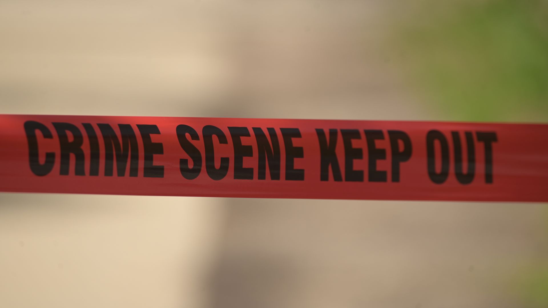 Red crime-scene tape with black letters reading "CRIME SCENE KEEP OUT" across the bottom; blurred beige pathway and green foliage in the background.