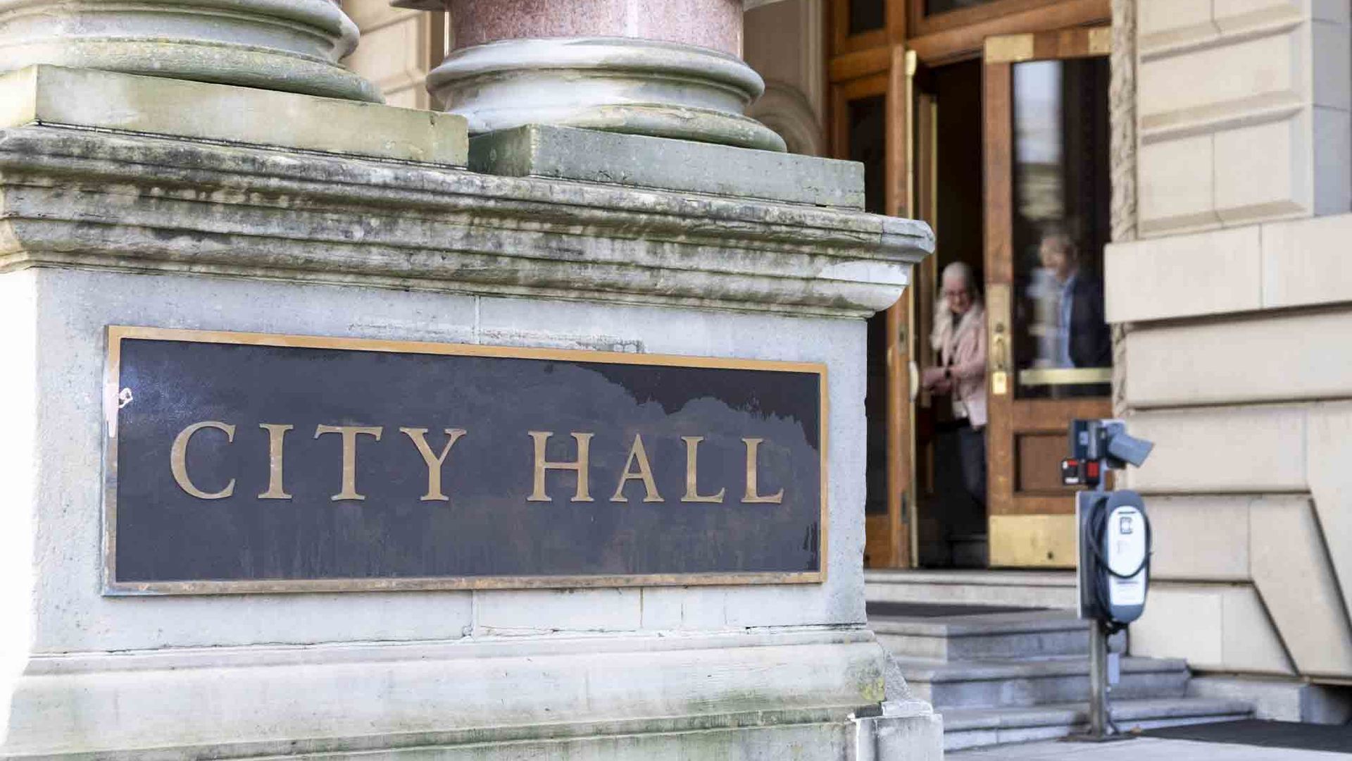 Close-up of a city hall facade featuring pink granite columns with round bases, a dark plaque that reads CITY HALL, stone steps, and a doorway with wooden frames; a few people visible in the background.