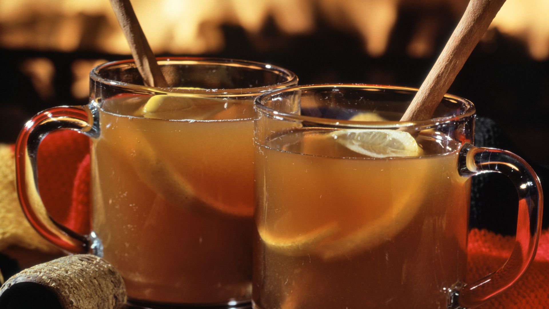 Two glass cups hot apple cider with cinnamon sticks and a roaring fireplace in the background.