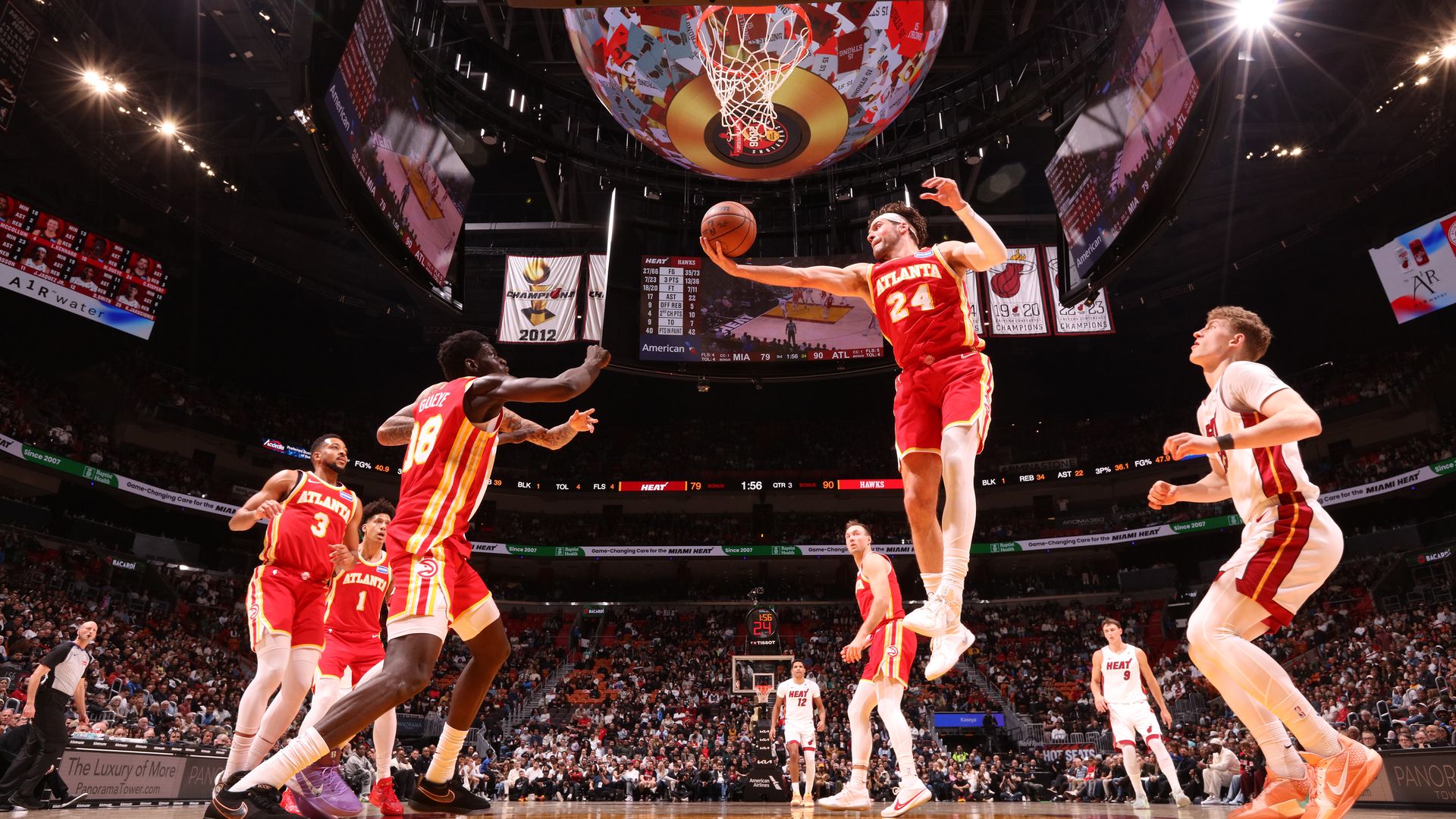 Basketball players wearing red and white uniforms watch as one player catches a rebound from a shot. 