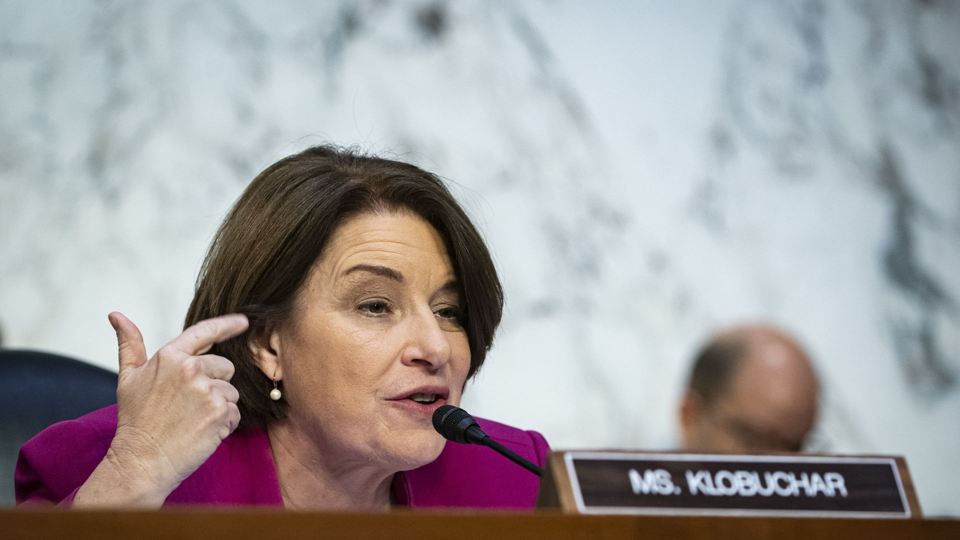 A person speaks in front of a microphone and a nameplate that says "Ms. Klobuchar"