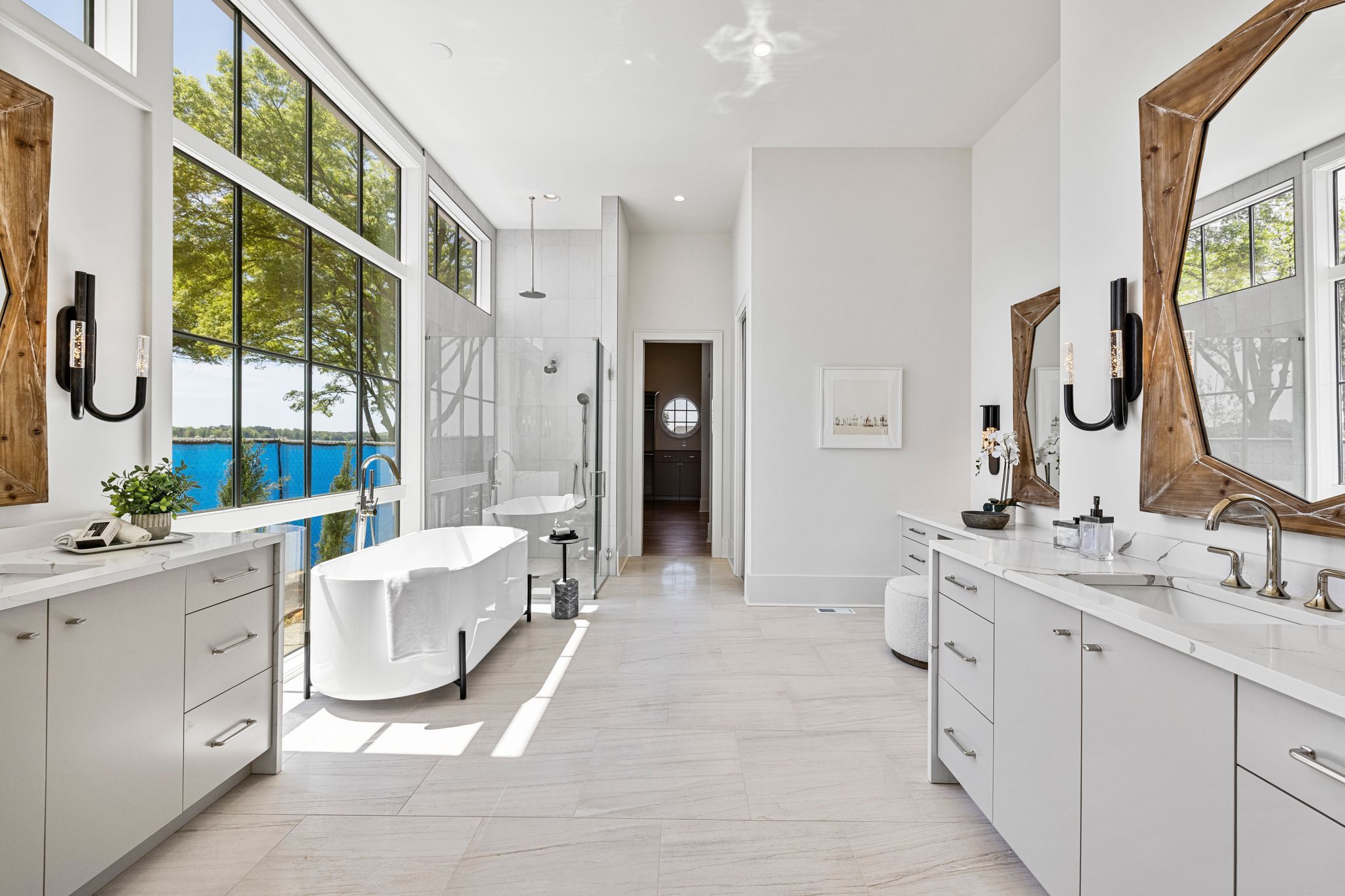 Bright modern bathroom with large windows overlooking trees and water, a white freestanding tub, glass shower, double sinks with wood-framed mirrors, light wood floors, and white walls.