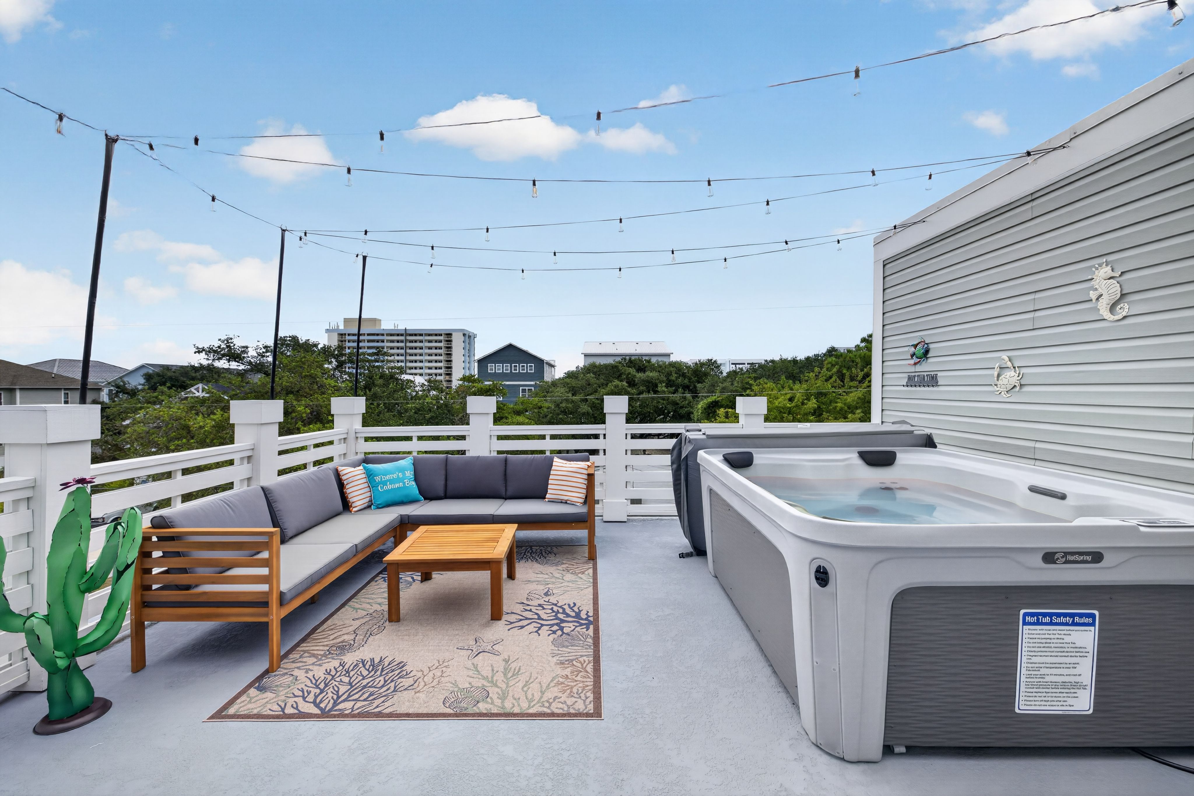 Rooftop deck with string lights overhead, a gray outdoor sectional with orange and teal pillows around a wooden coffee table, a sea-life rug, white railing, and a large hot tub.