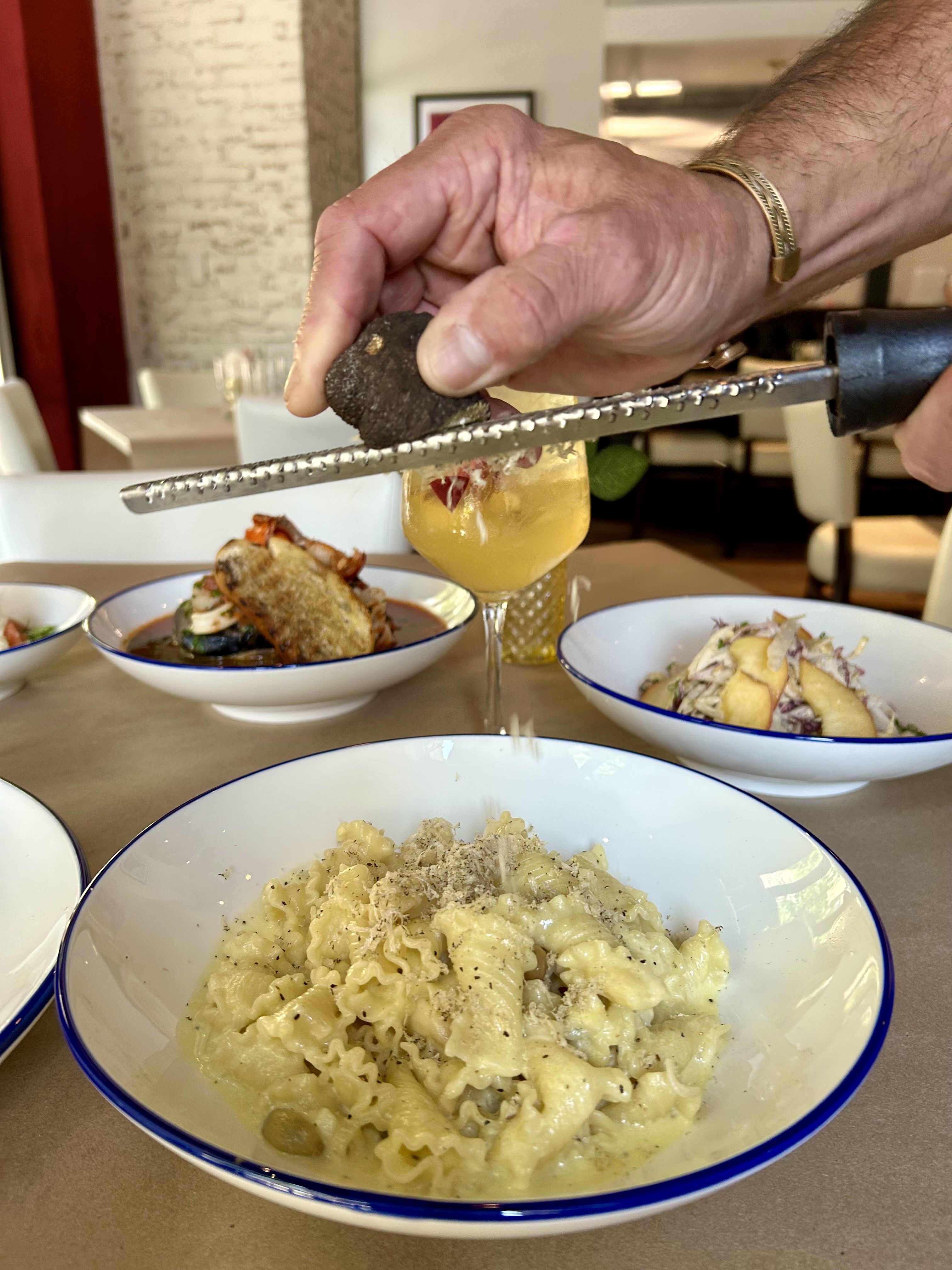 Hand grating black truffle over creamy pasta in a white bowl with a blue rim, with dishes and a yellow drink on a restaurant table in the background.