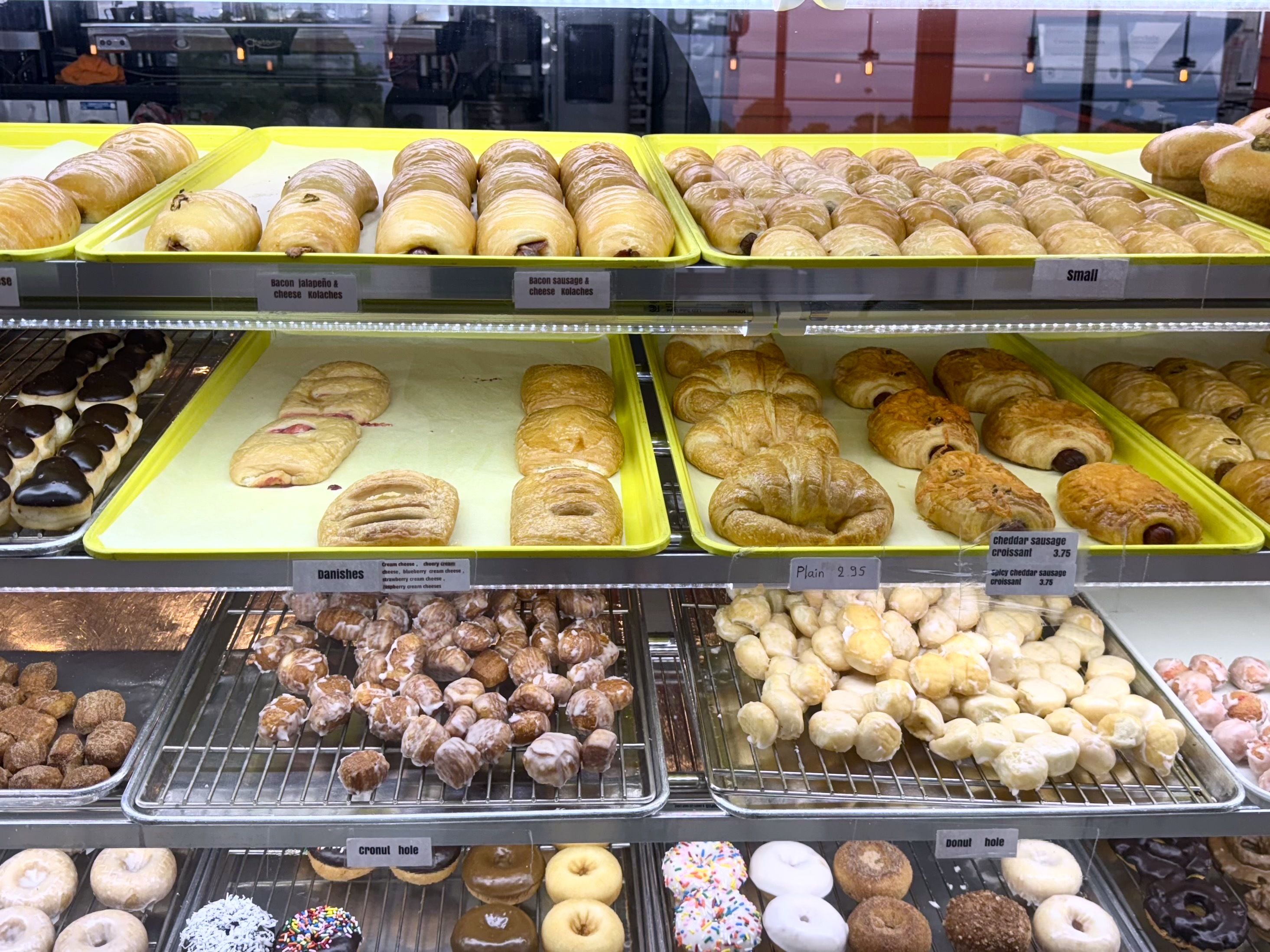Bakery display case filled with kolaches, croissants, donut holes, and pastries on yellow trays and metal racks.